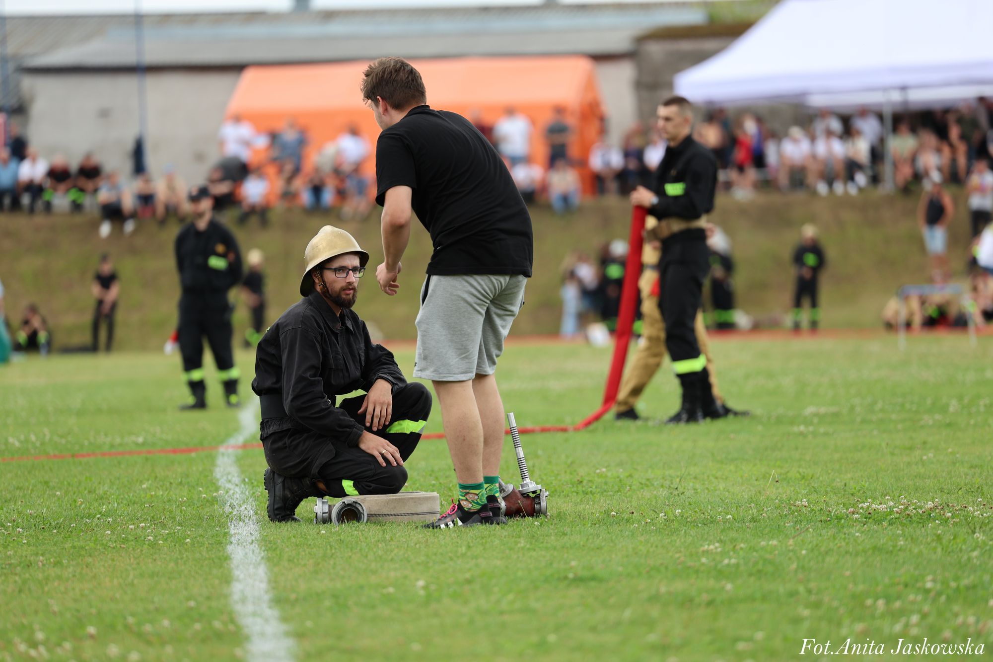 Mężczyzna w czarnym ubraniu strażackim i hełmie kuca na trawie obok mężczyzny w czarnej koszulce i szarych spodenkach, w tle rozmyta grupa ludzi i biały namiot.