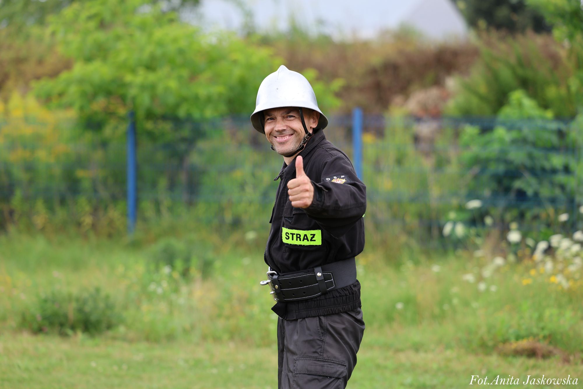Mężczyzna w czarnym mundurze strażackim z zieloną naszywką "STRAŻ" i srebrnym hełmem, pokazujący kciuk w górę na tle zieleni i ogrodzenia.