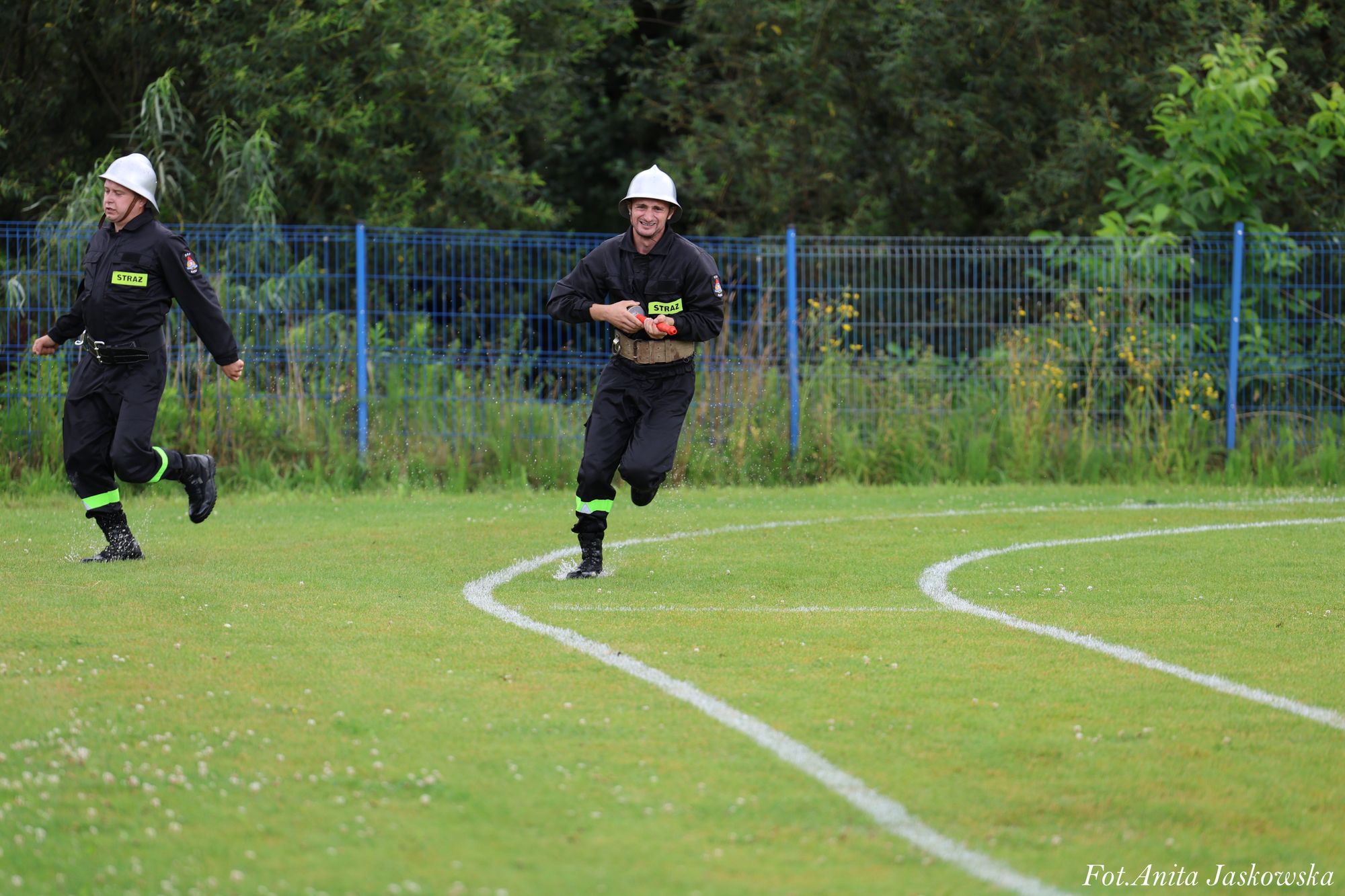 Dwóch strażaków w czarnych mundurach i białych hełmach biegnie po zielonym boisku sportowym z białymi liniami, w tle niebieski metalowy płot i zarośla.