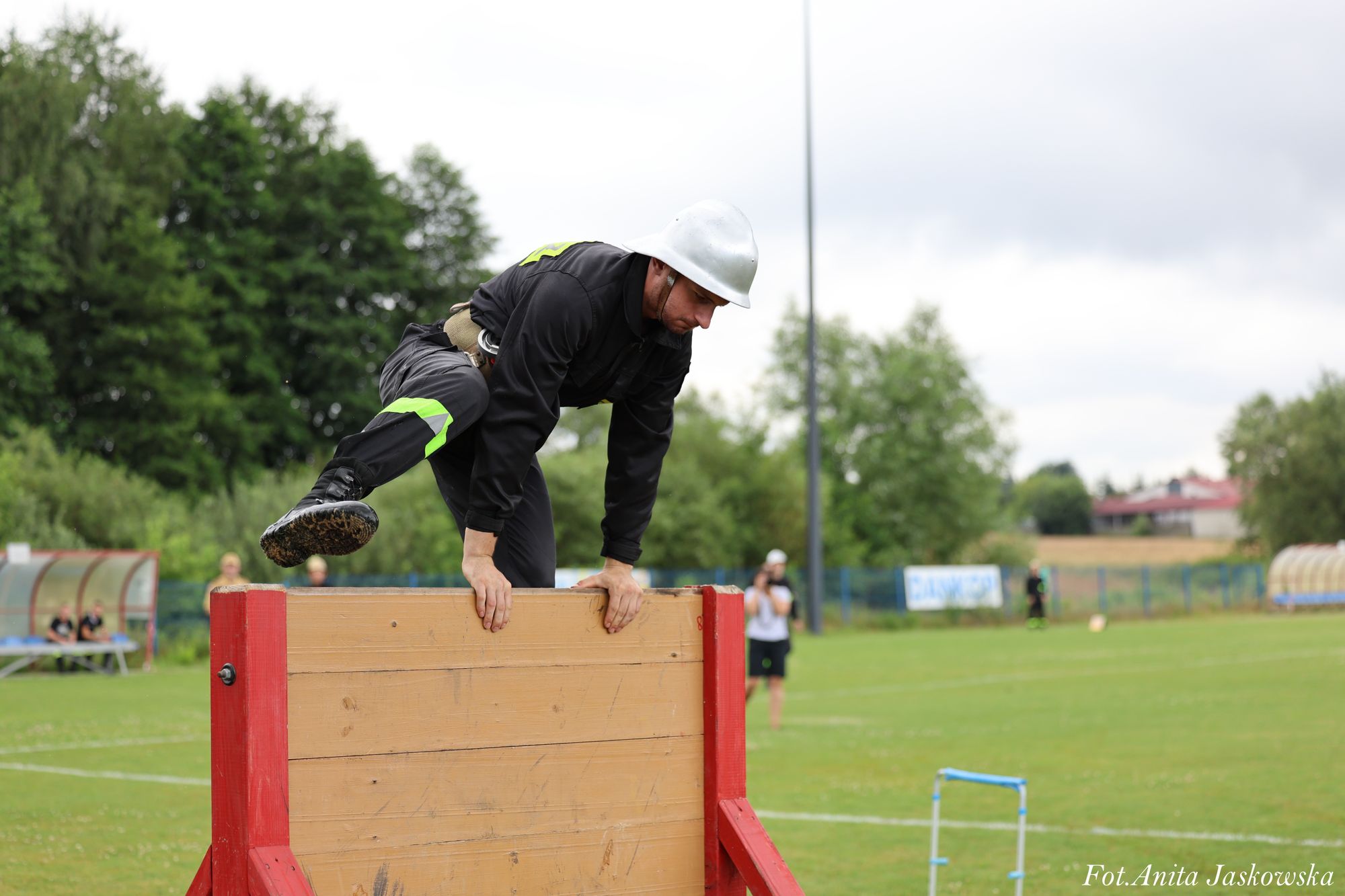 Mężczyzna w czarnym ubraniu i białym kasku pokonuje drewnianą przeszkodę na zielonym boisku sportowym, w tle drzewa i osoby na ławce oraz fotograf.