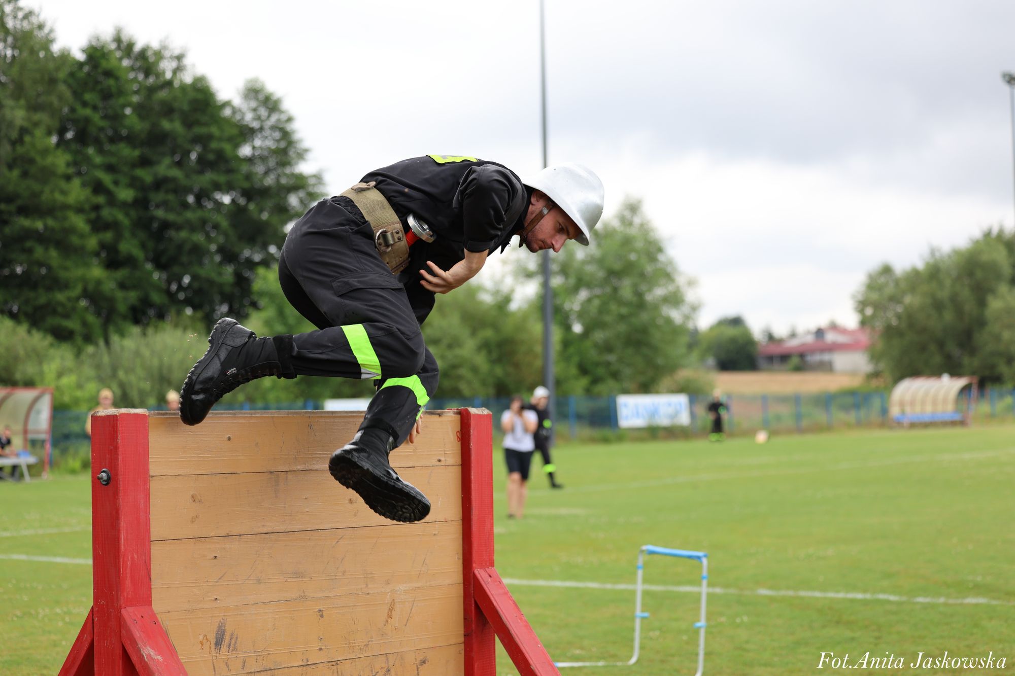 Mężczyzna w czarnym ubraniu z odblaskowymi zielonymi paskami i białym hełmem skacze przez drewnianą przeszkodę na zielonym boisku, w tle drzewa i budynki.