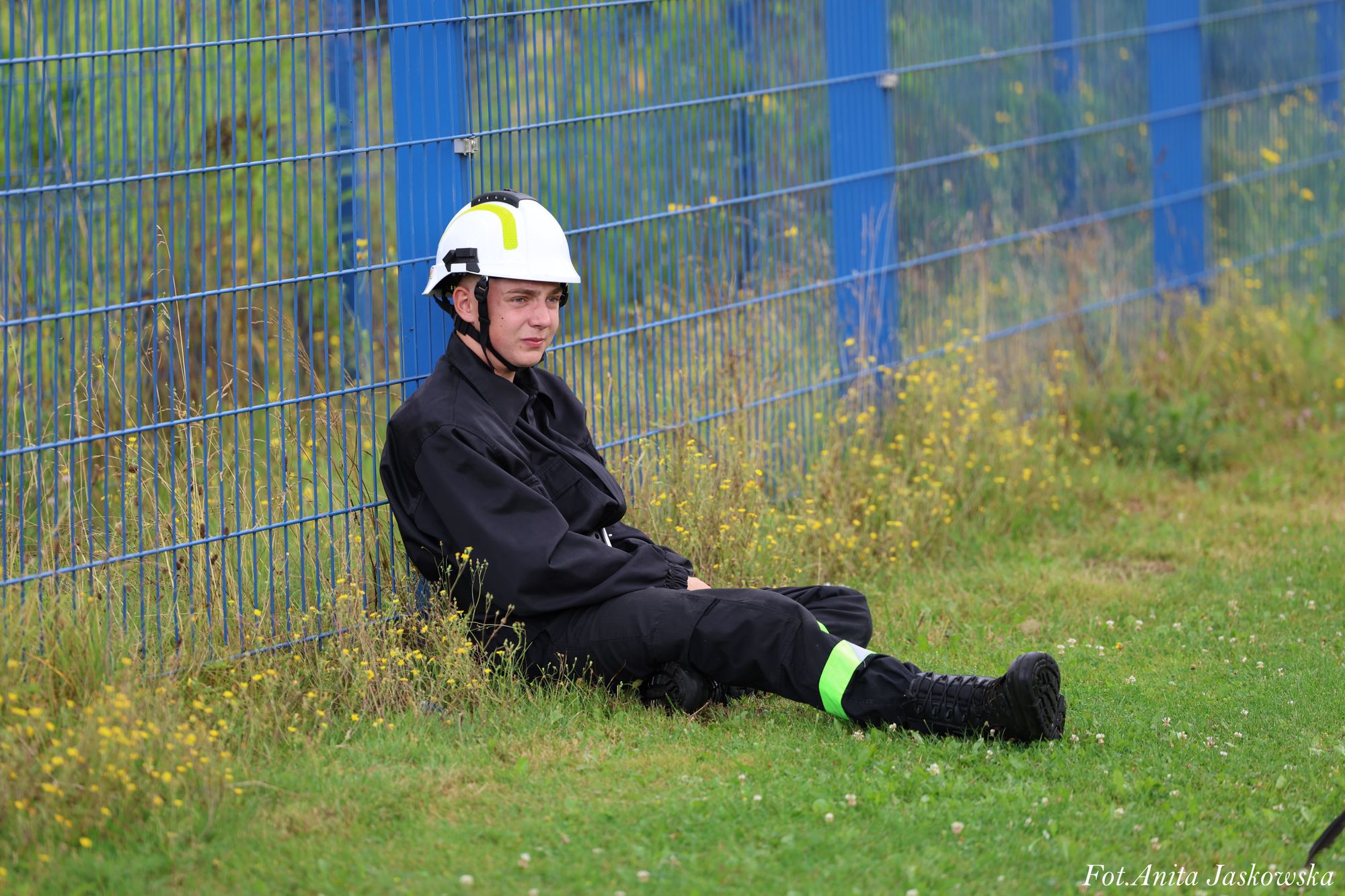 Młody mężczyzna w czarnym ubraniu roboczym z odblaskowym paskiem i białym kaskiem siedzi na trawie oparty o niebieski metalowy płot, wokół żółte kwiaty.