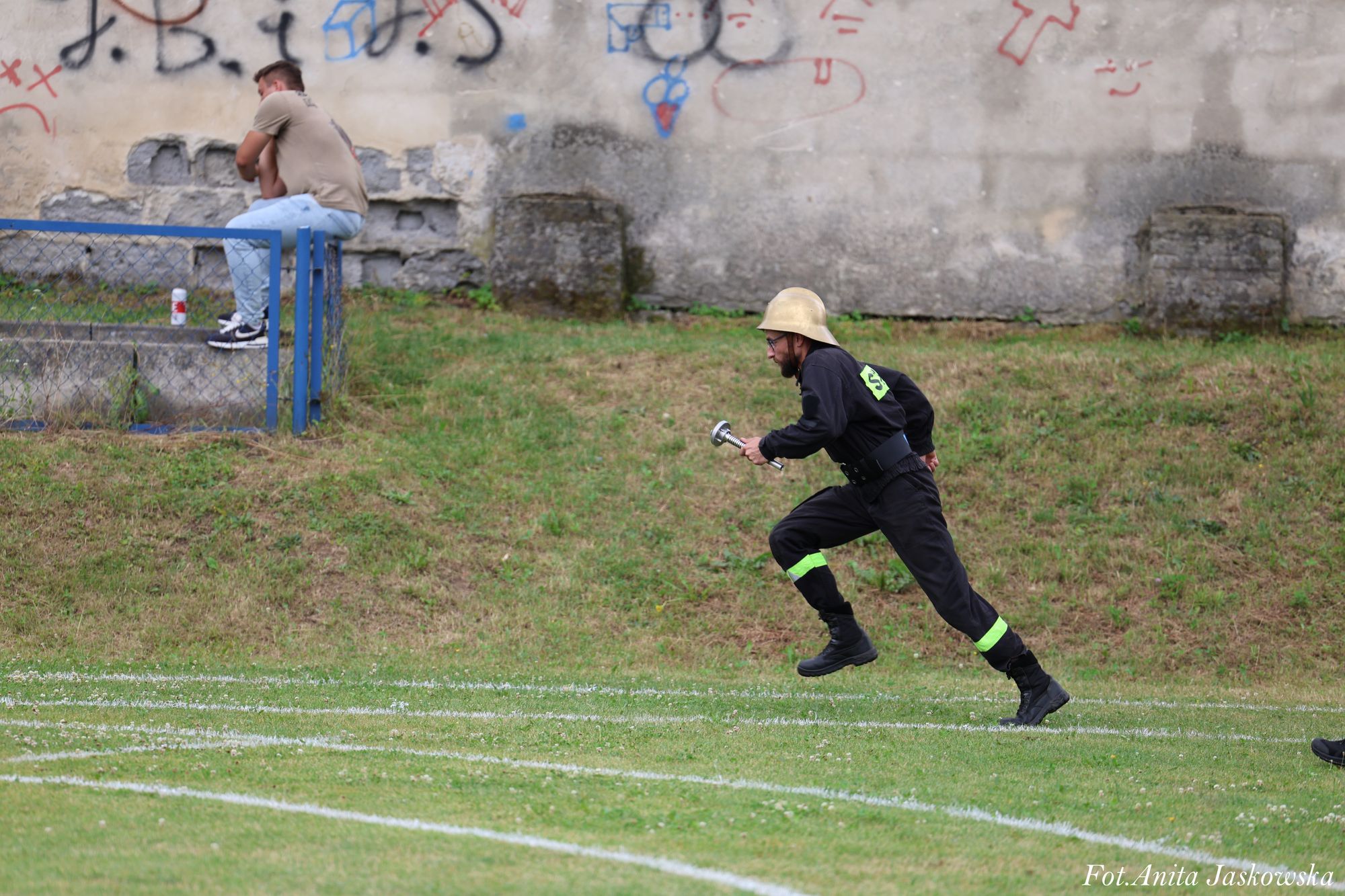 Mężczyzna w czarnym uniformie z odblaskowymi paskami i hełmem w biegu po zielonym boisku z białymi liniami, w tle mężczyzna siedzący na murku z niebieską barierką i ściana z graffiti.