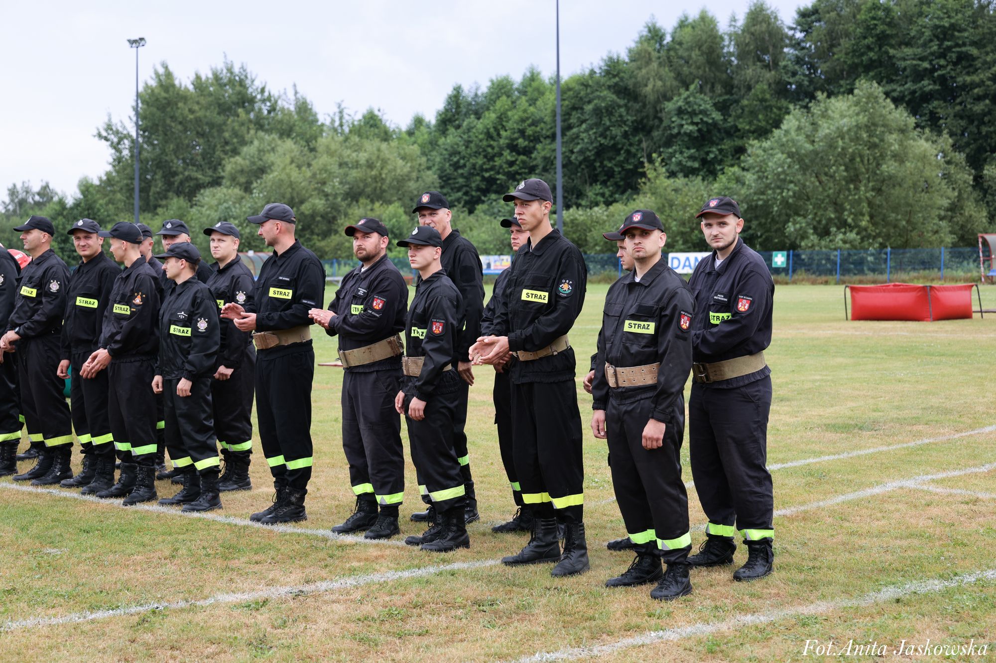Grupa mężczyzn w czarnych mundurach z odblaskowymi paskami i napisami "STRAŻ" stoi na zielonym boisku sportowym, w tle drzewa i niebo.