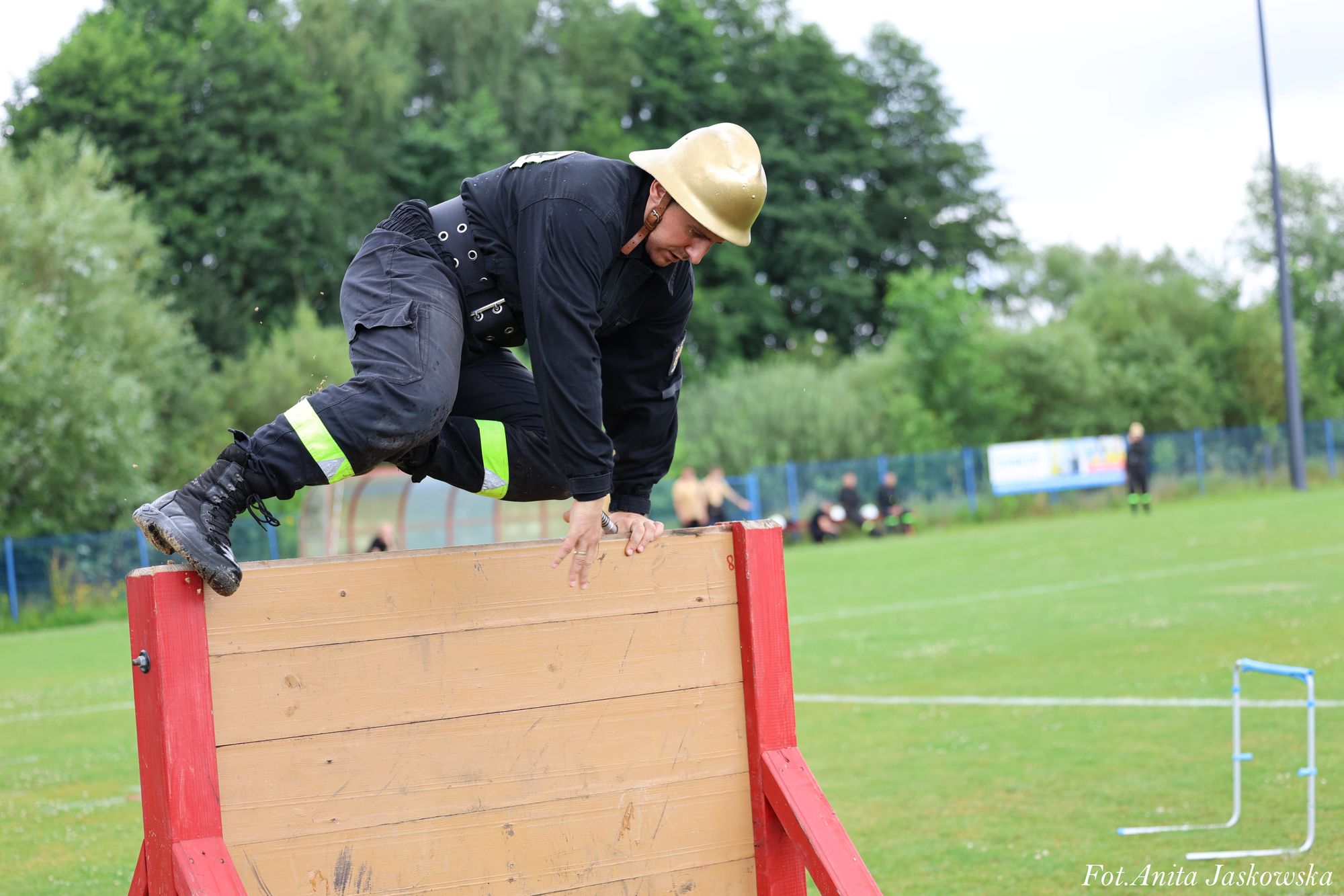 Mężczyzna w czarnym stroju i żółtym hełmie pokonuje drewnianą przeszkodę na zielonym boisku z drzewami w tle.