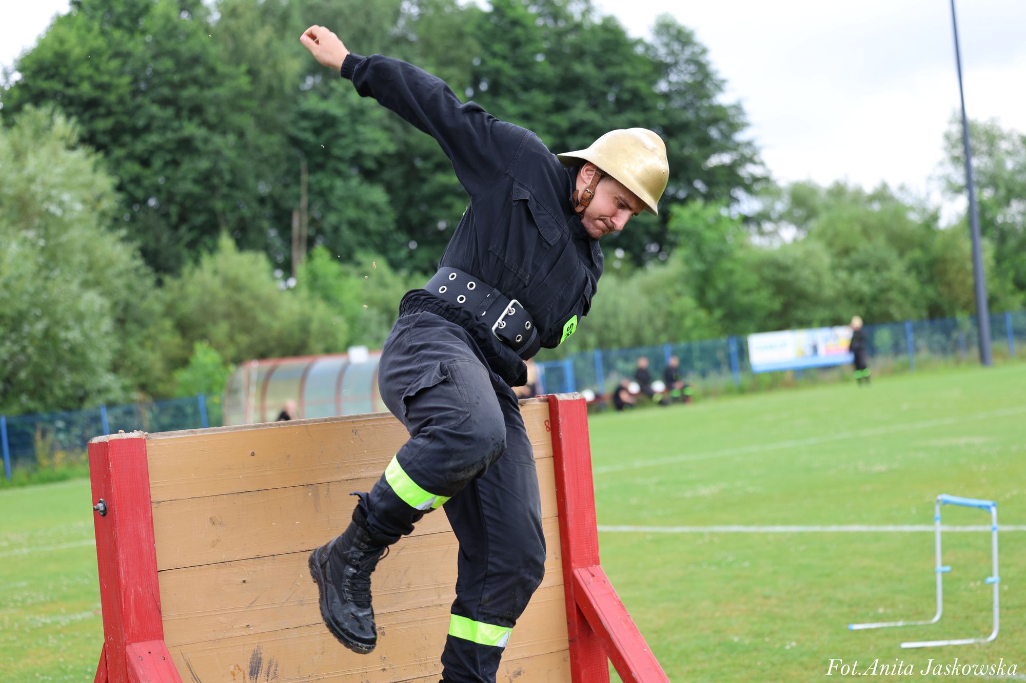 Mężczyzna w czarnym ubraniu i złotym hełmie skacze przez drewnianą przeszkodę na zielonym boisku z drzewami w tle.