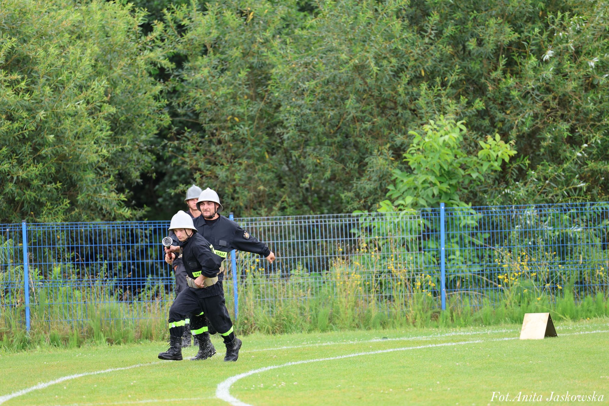 Trzech mężczyzn w czarnych mundurach i srebrnych hełmach na zielonym boisku z niebieskim płotem i gęstą zieloną roślinnością w tle.