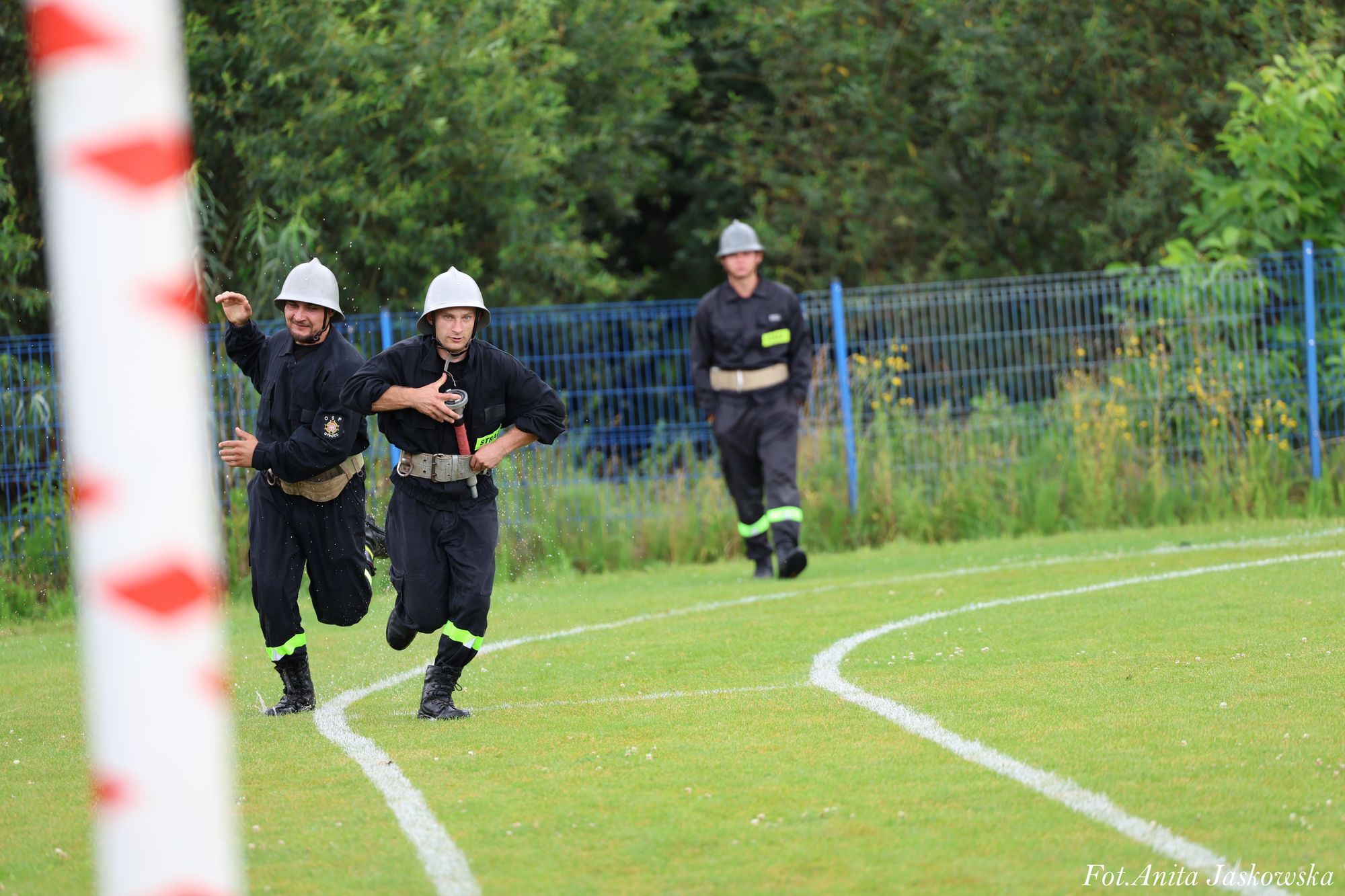 Trzech mężczyzn w czarnych mundurach strażackich i srebrnych hełmach biegnie po zielonej trawie z białymi liniami, w tle niebieski płot i drzewa.