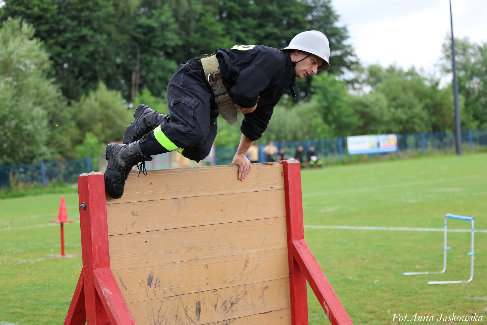 Mężczyzna w czarnym umundurowaniu i białym hełmie pokonuje przeszkodę z jasnobrązowej deski z czerwonymi słupkami na zielonym polu.