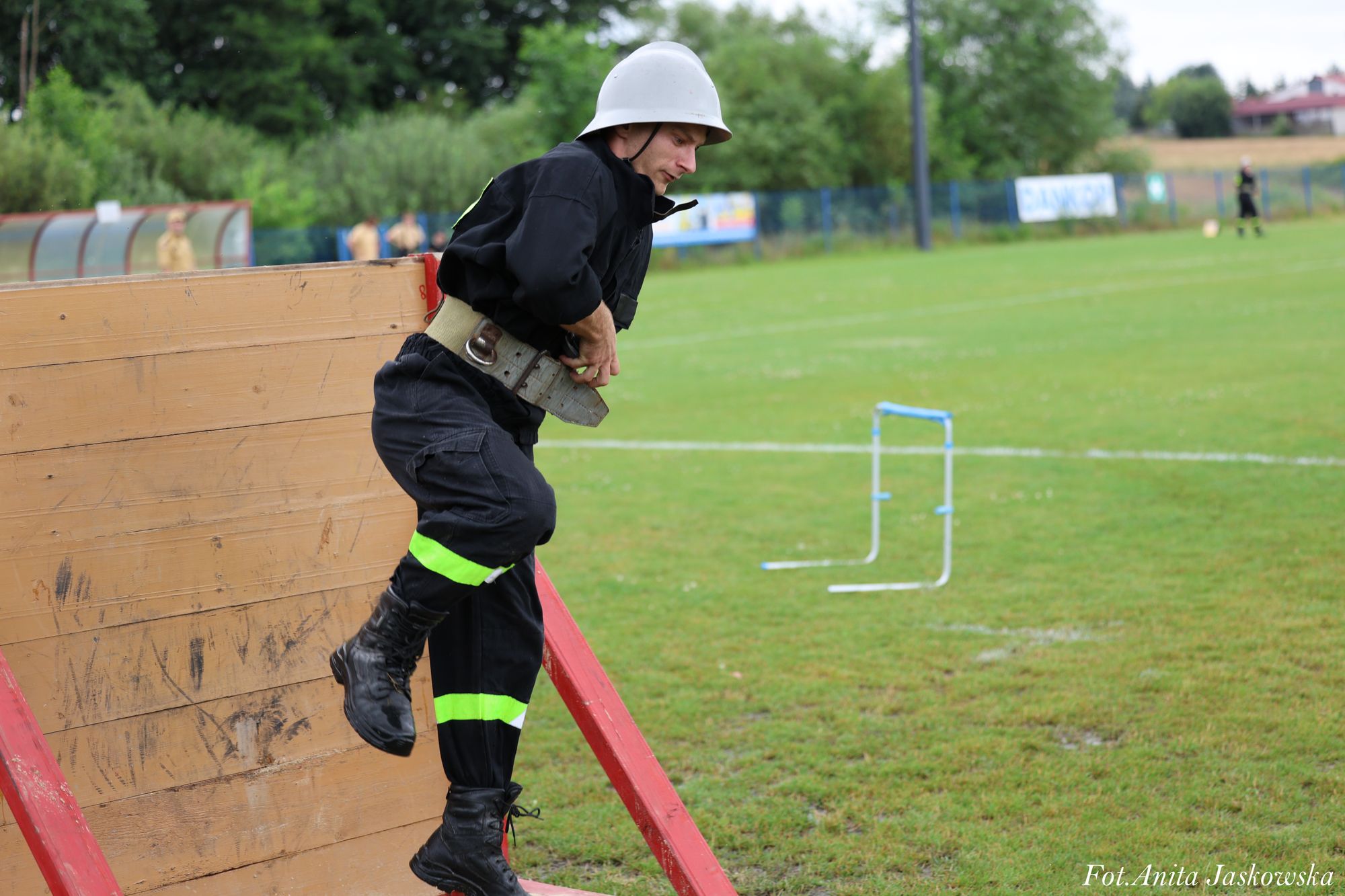 Mężczyzna w czarnym stroju strażaka z białym hełmem pokonuje drewnianą przeszkodę na zielonym boisku sportowym.