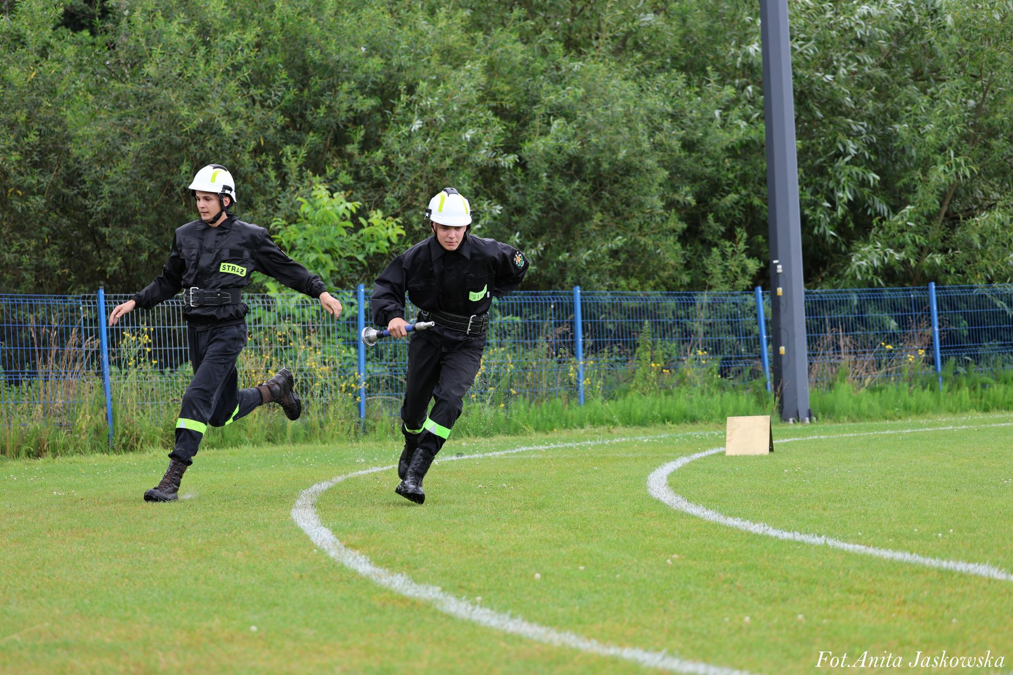 Dwóch strażaków w czarnych uniformach i białych hełmach biegnie po zielonym boisku sportowym z białymi liniami, w tle niebieski płot i zarośla.