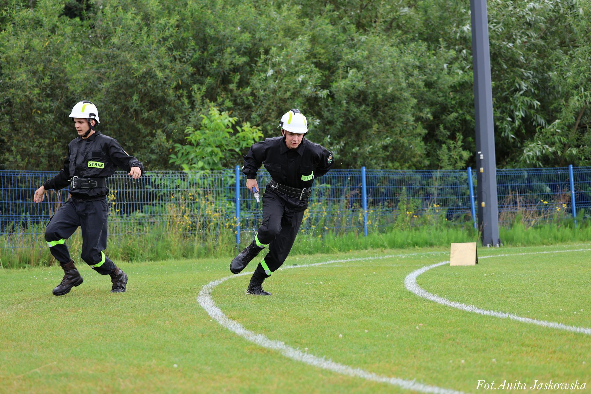 Dwóch mężczyzn w czarnych mundurach i białych hełmach biegnie po zielonym boisku z białymi liniami, w tle niebieski metalowy płot i gęste zielone krzewy.