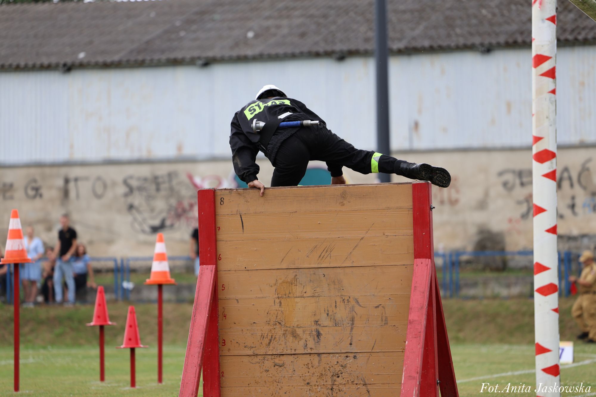 Strażak w czarnym ubraniu i białym kasku pokonuje drewnianą przeszkodę z czerwonymi krawędziami na zielonym boisku z widocznymi pachołkami i metalową bramką.