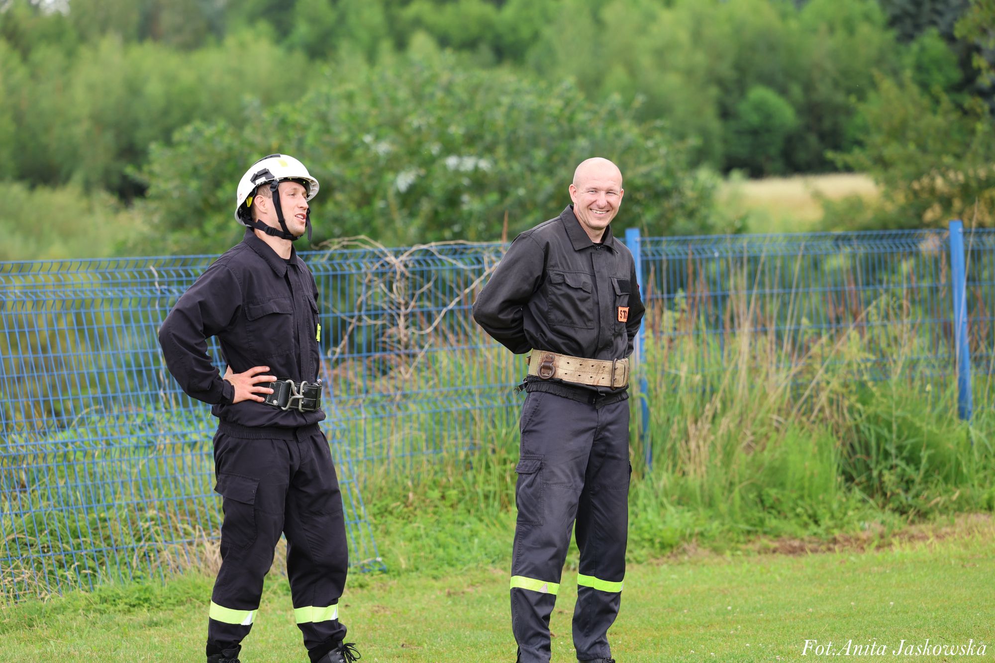Dwóch mężczyzn w czarnych mundurach strażackich z odblaskowymi pasami, jeden w białym hełmie, stoją na trawie przy niebieskim ogrodzeniu, w tle zielone krzewy.