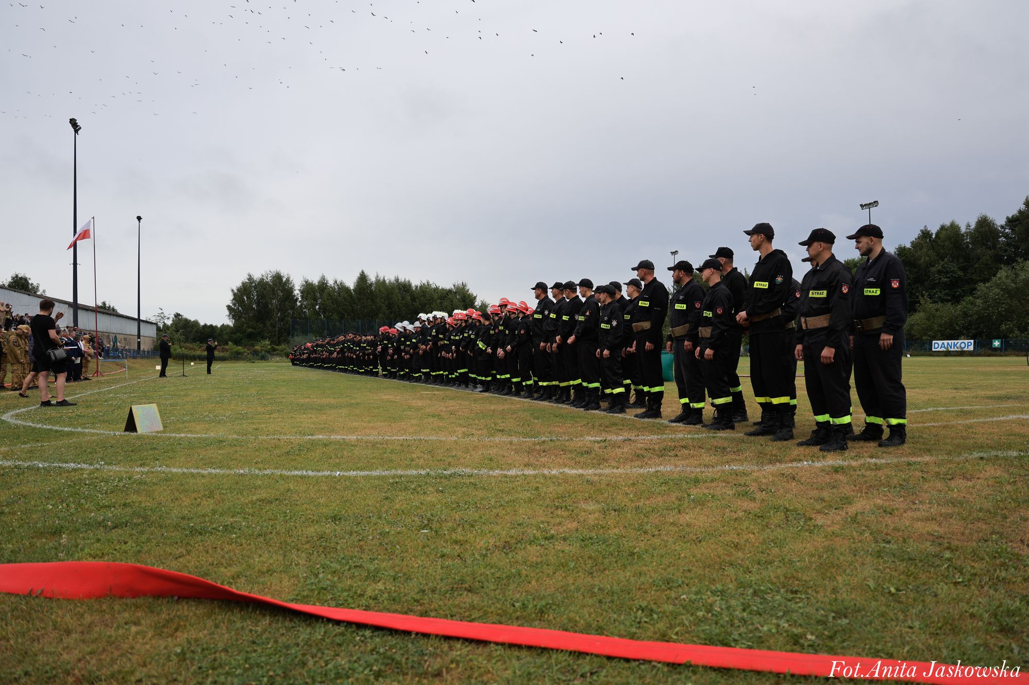 Rząd strażaków w czarnych mundurach z żółtymi odblaskami i kaskami, ustawionych na zielonym boisku pod pochmurnym niebem, po lewej stronie widoczna flaga Polski.