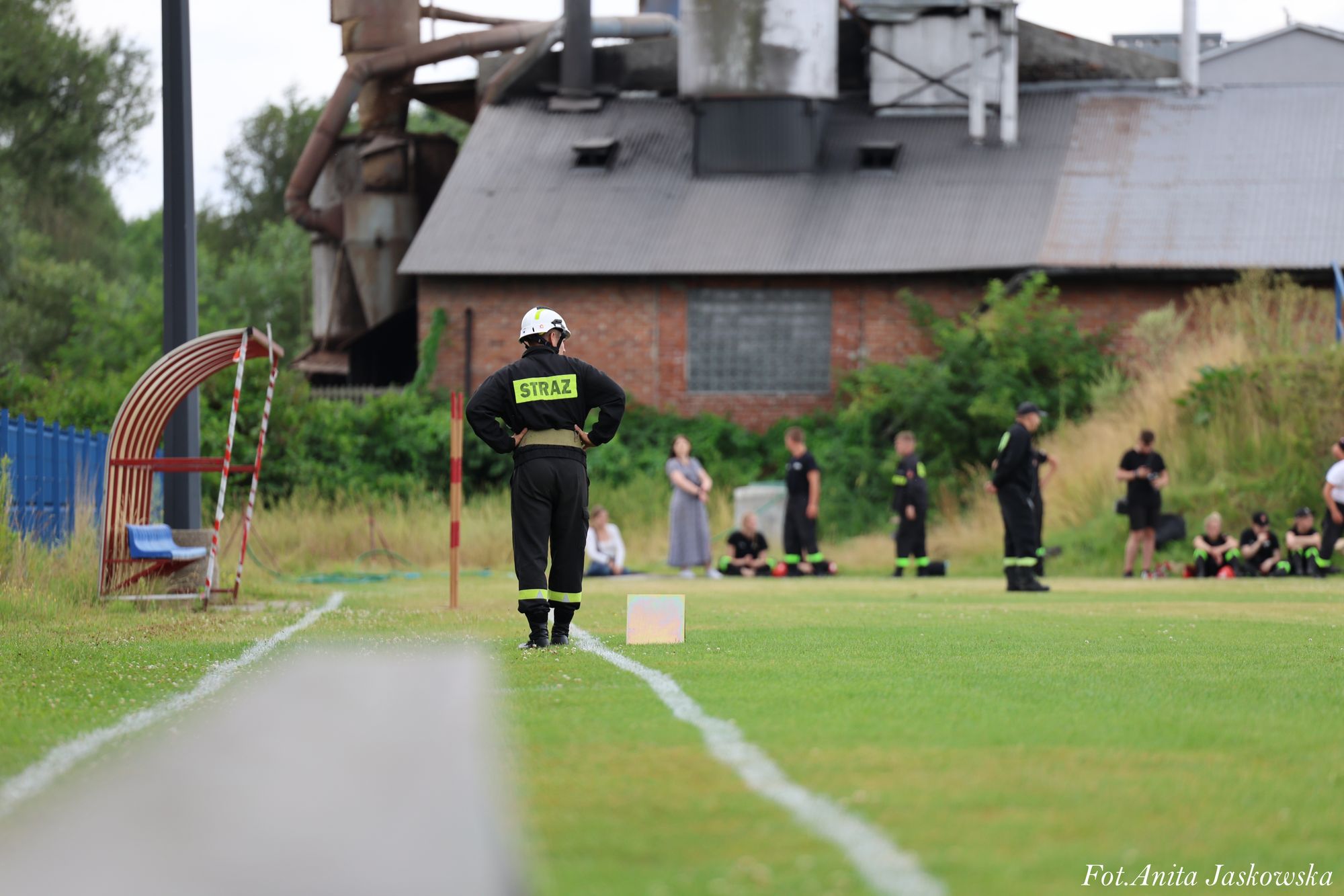 Strażak w czarnym mundurze z napisem "STRAŻ" na plecach stoi na zielonym boisku piłkarskim, w tle budynek i grupa osób w mundurach.