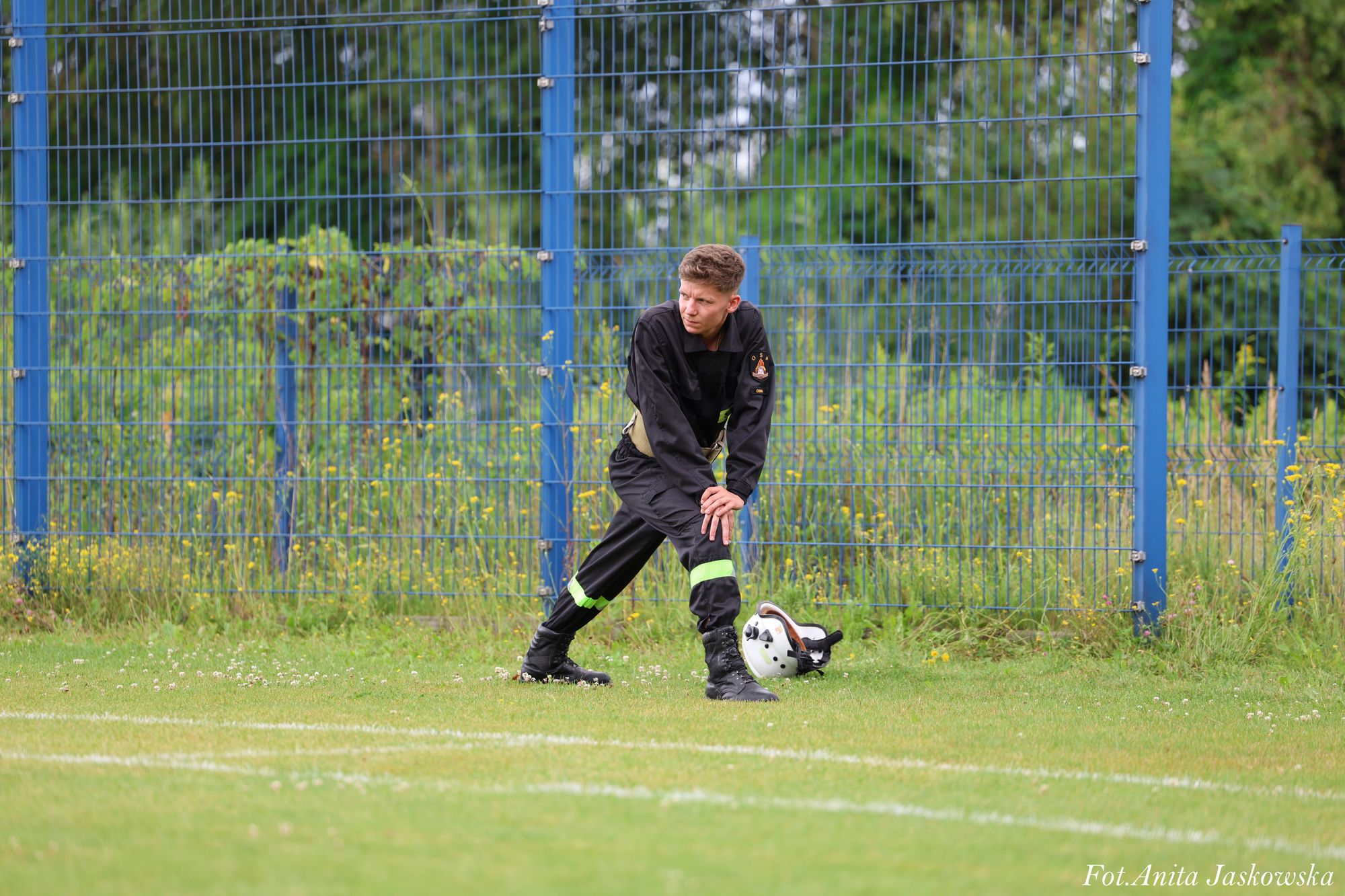 Młody strażak w czarnym ubraniu z odblaskowymi paskami rozciąga nogi na trawie obok białego hełmu, za nim niebieski metalowy płot i zieleń.