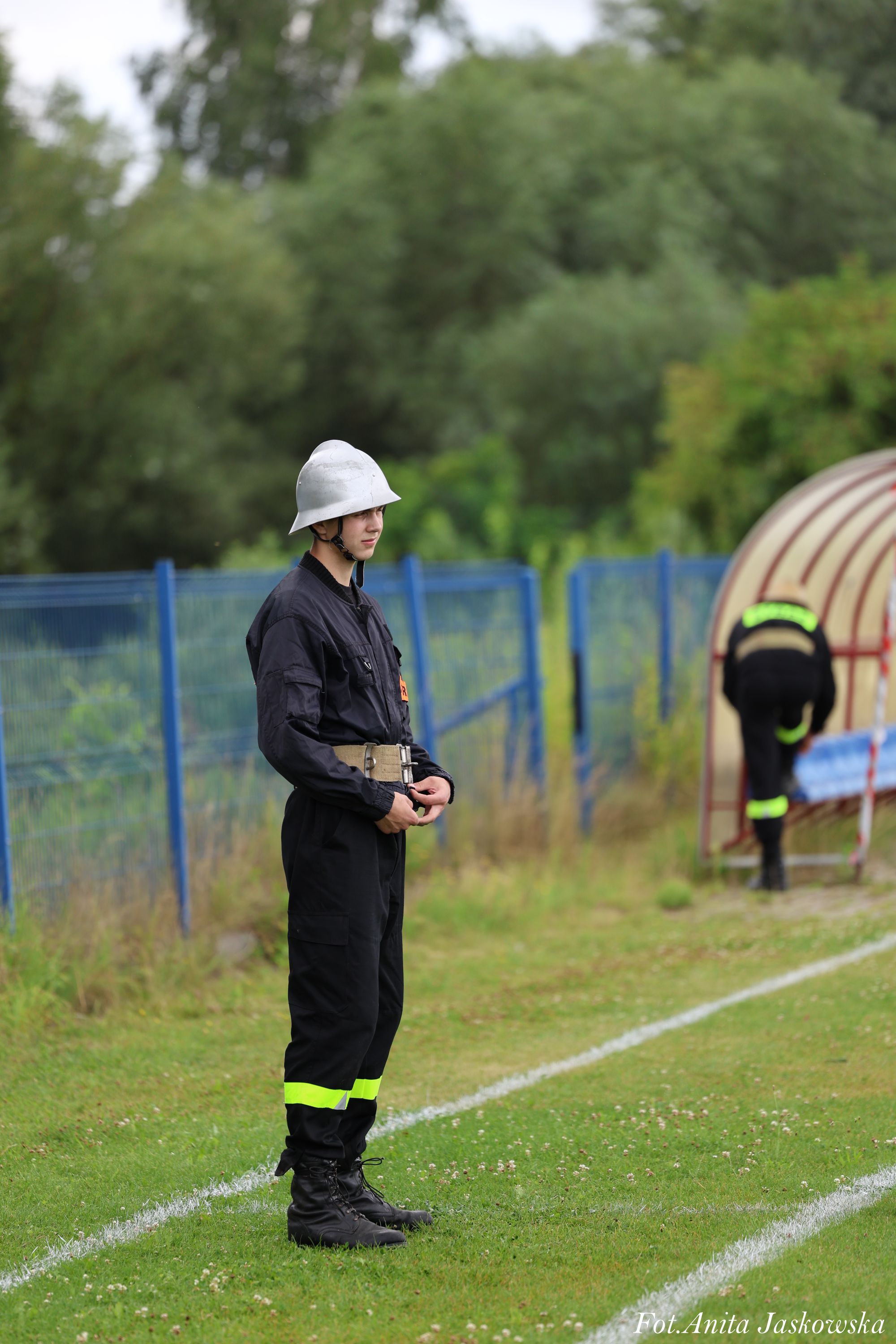 Młody strażak w czarnym mundurze z żółtymi odblaskami i białym hełmem stoi na zielonej trawie obok niebieskiego ogrodzenia, w tle druga osoba przy czerwonym tunelu.