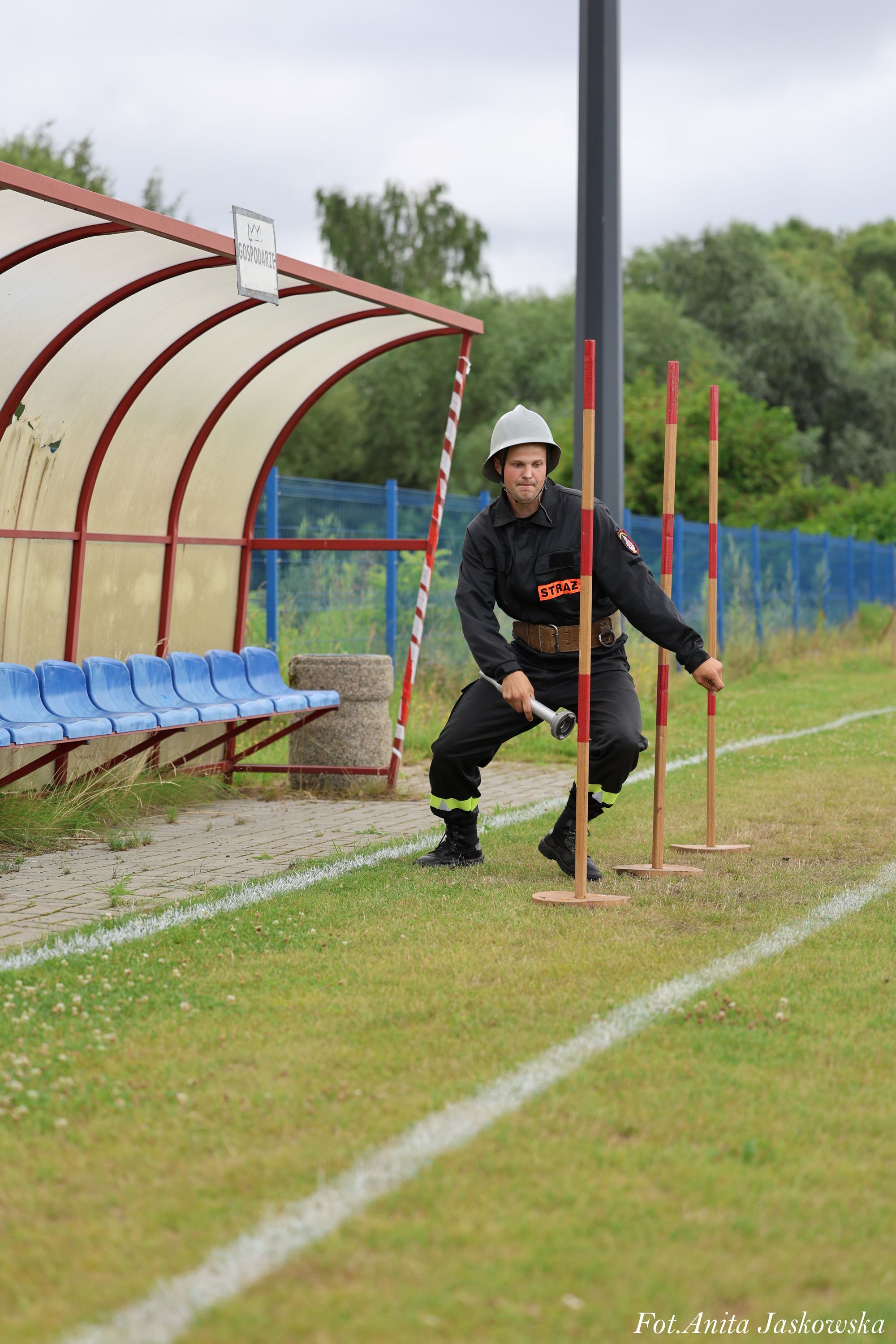 Strażak w czarnym mundurze i hełmie ćwiczy na trawie między trzema pionowymi drewnianymi tyczkami, obok niebieskich siedzeń pod zadaszeniem.