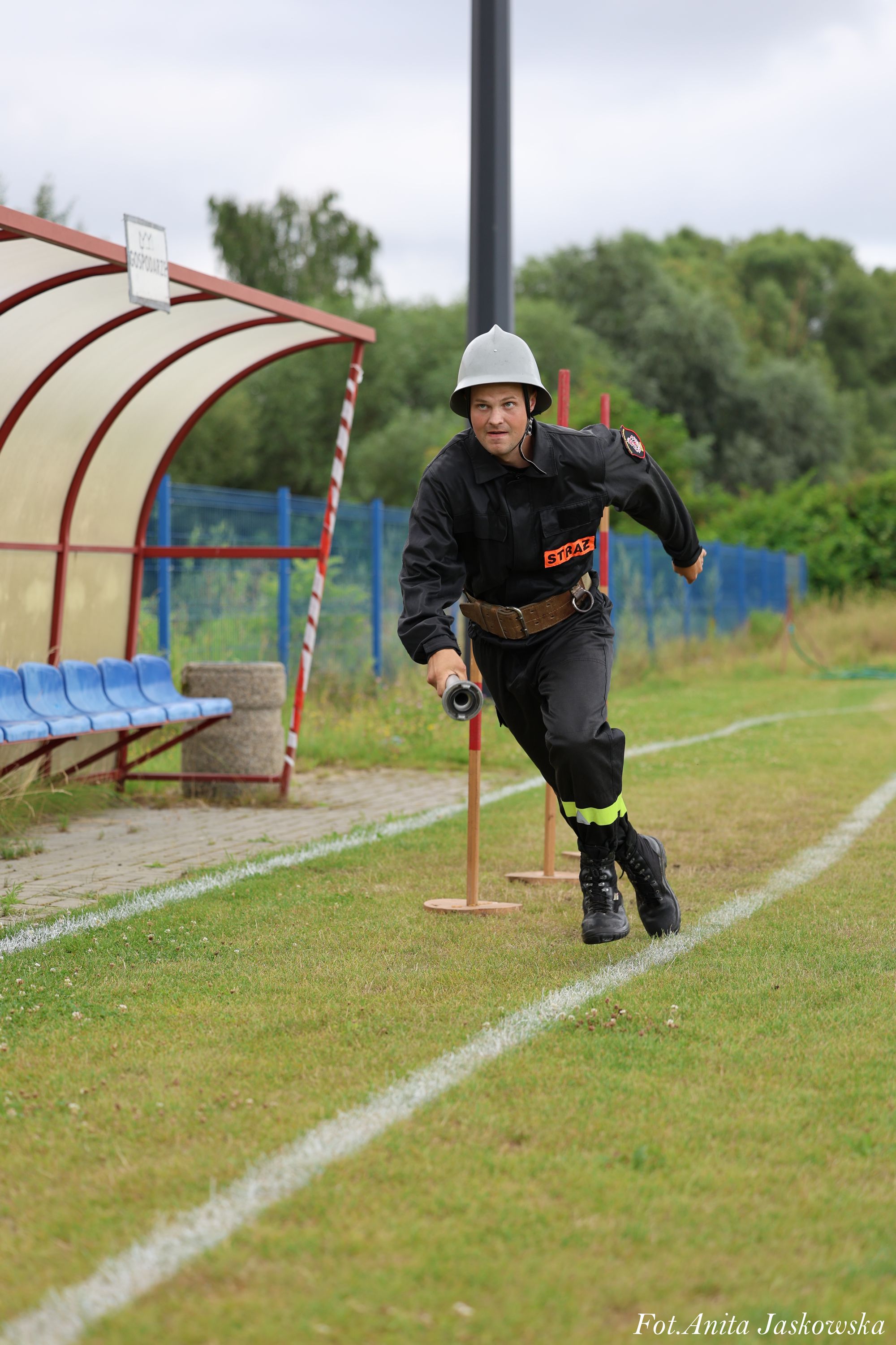 Mężczyzna w czarnym mundurze strażackim i szarym hełmie biegnie po trawiastej nawierzchni obok niebieskich siedzeń pod zadaszeniem, trzyma metalowe narzędzie.