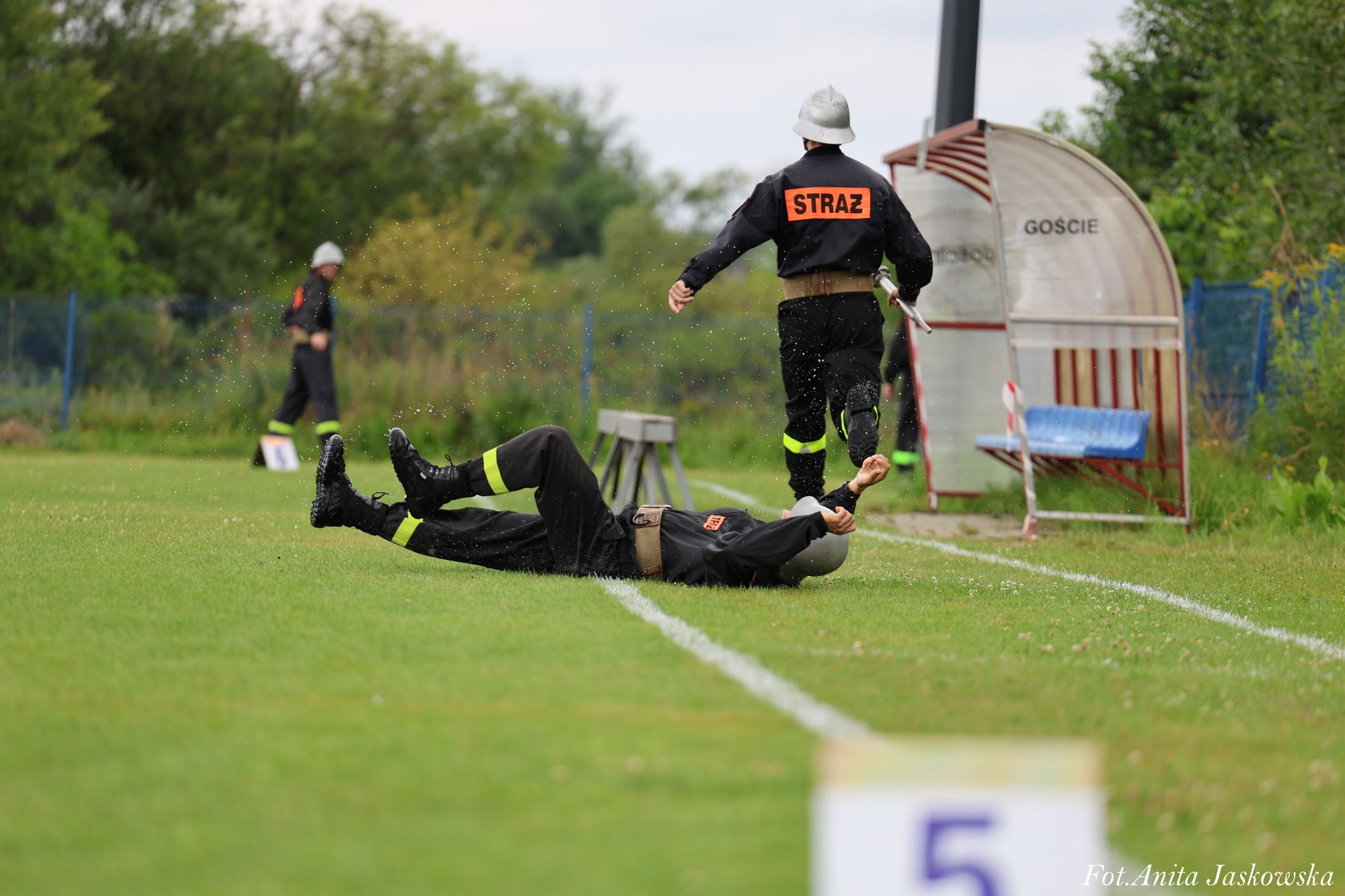 Dwóch strażaków w czarnych mundurach z żółtymi lampasami, jeden leży na trawie, drugi idzie w stronę zadaszonej ławki z napisem "GOŚCIE".