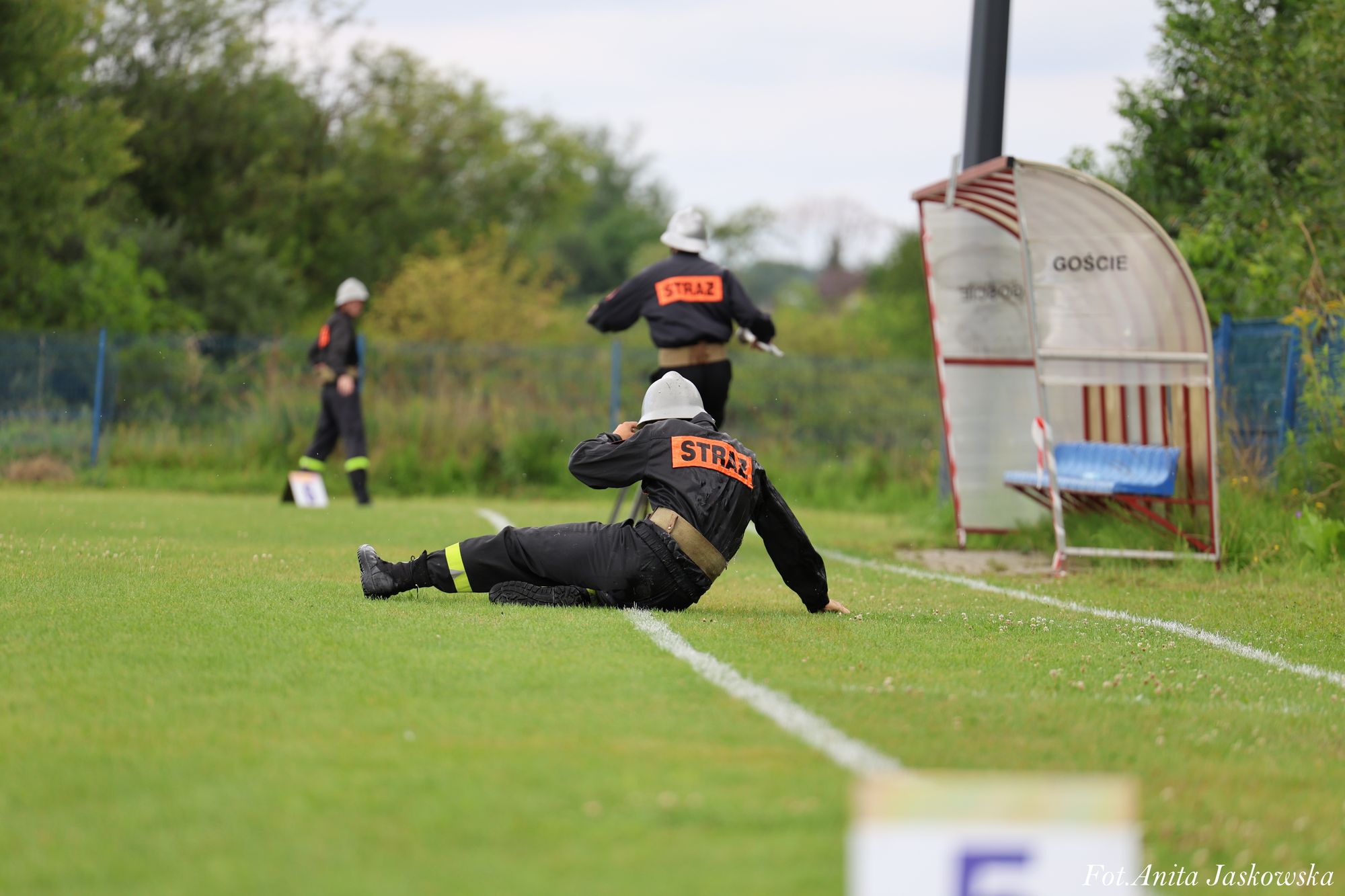 Trzy osoby w czarnych mundurach z napisem STRAŻ siedzą i stoją na zielonym boisku z białymi liniami, obok metalowa wiata z niebieskimi siedzeniami i napisem GOŚCIE.