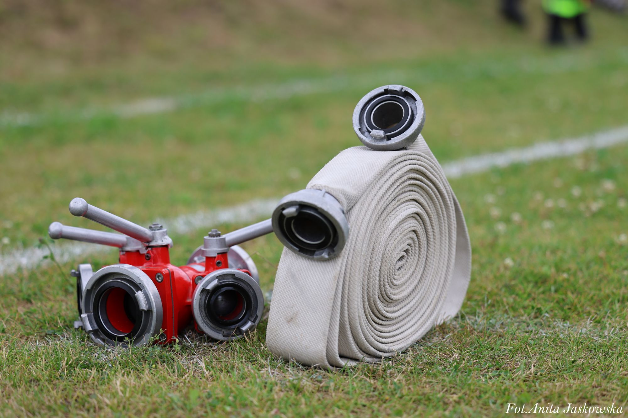 Biały zwinięty wąż strażacki z metalowymi końcówkami obok czerwonego rozdzielacza na zielonej trawie.