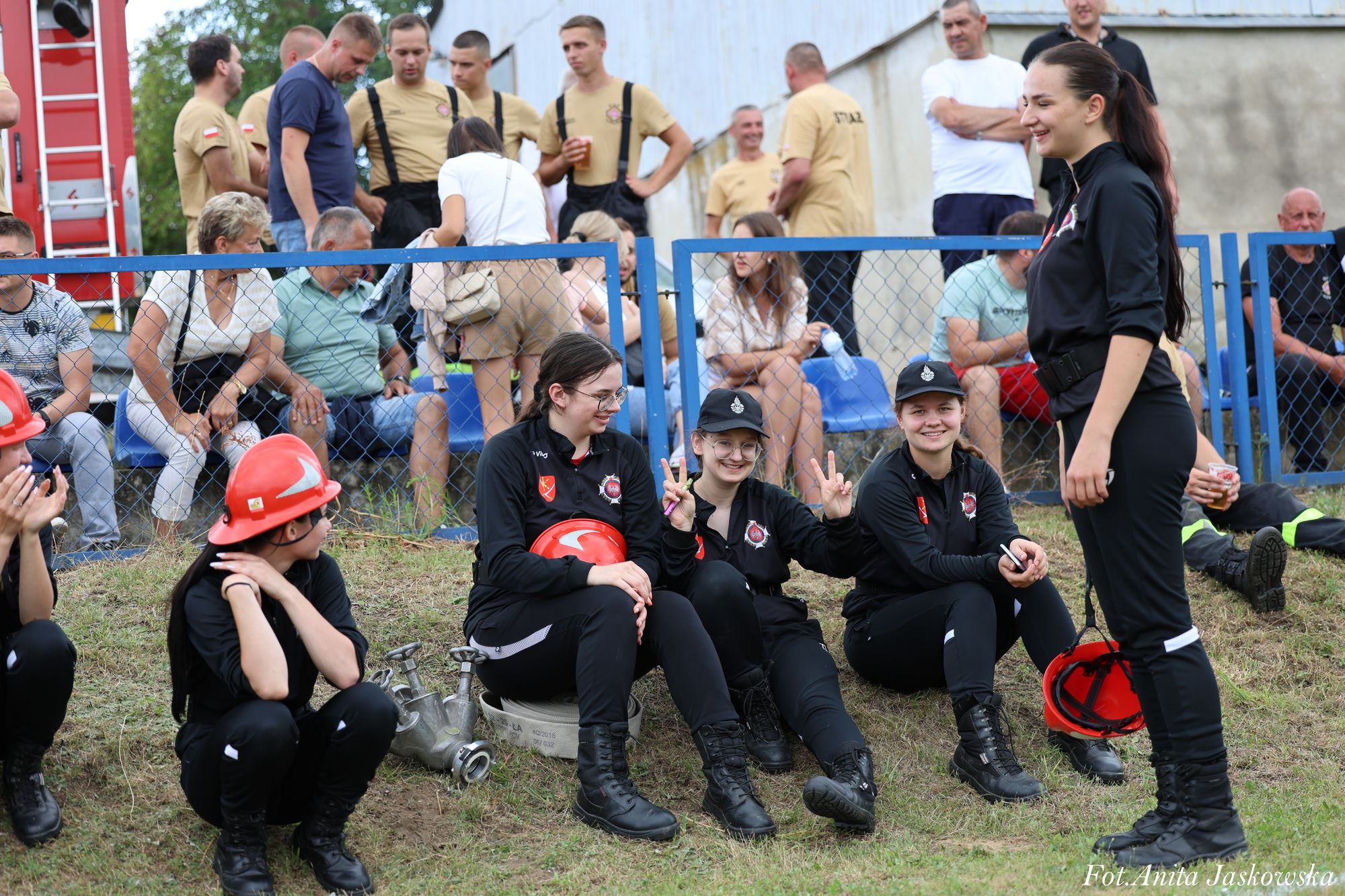 Grupa młodych osób w czarnych mundurach strażackich, niektóre z czerwonymi hełmami, siedząca i stojąca na trawie, za nimi ogrodzenie i ludzie na niebieskich krzesłach.