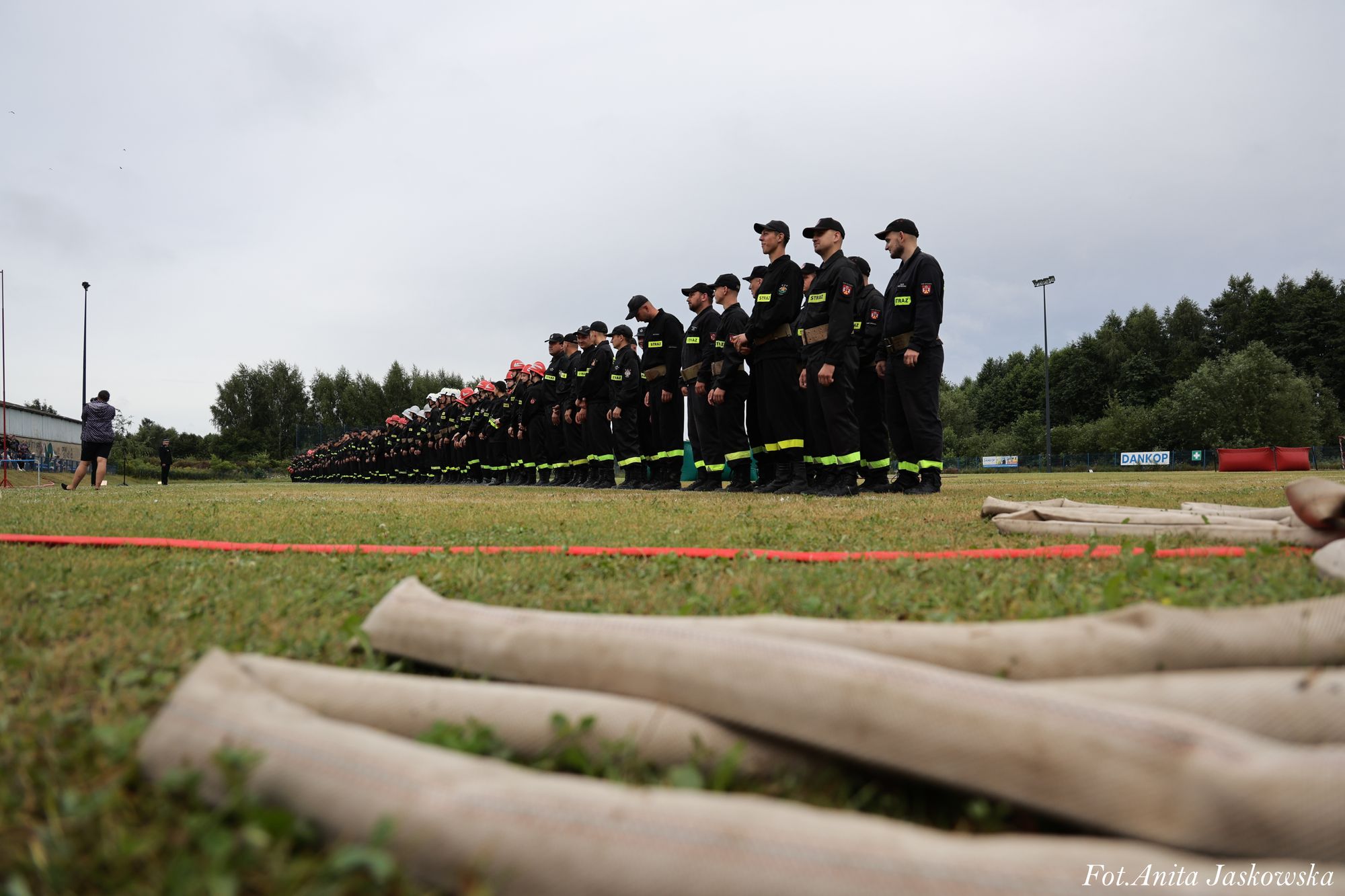 Szereg strażaków w czarnych mundurach z żółtymi pasami stoi na zielonym trawniku, na pierwszym planie zwinięte jasne węże strażackie, na tle zielonych drzew i szarego nieba.