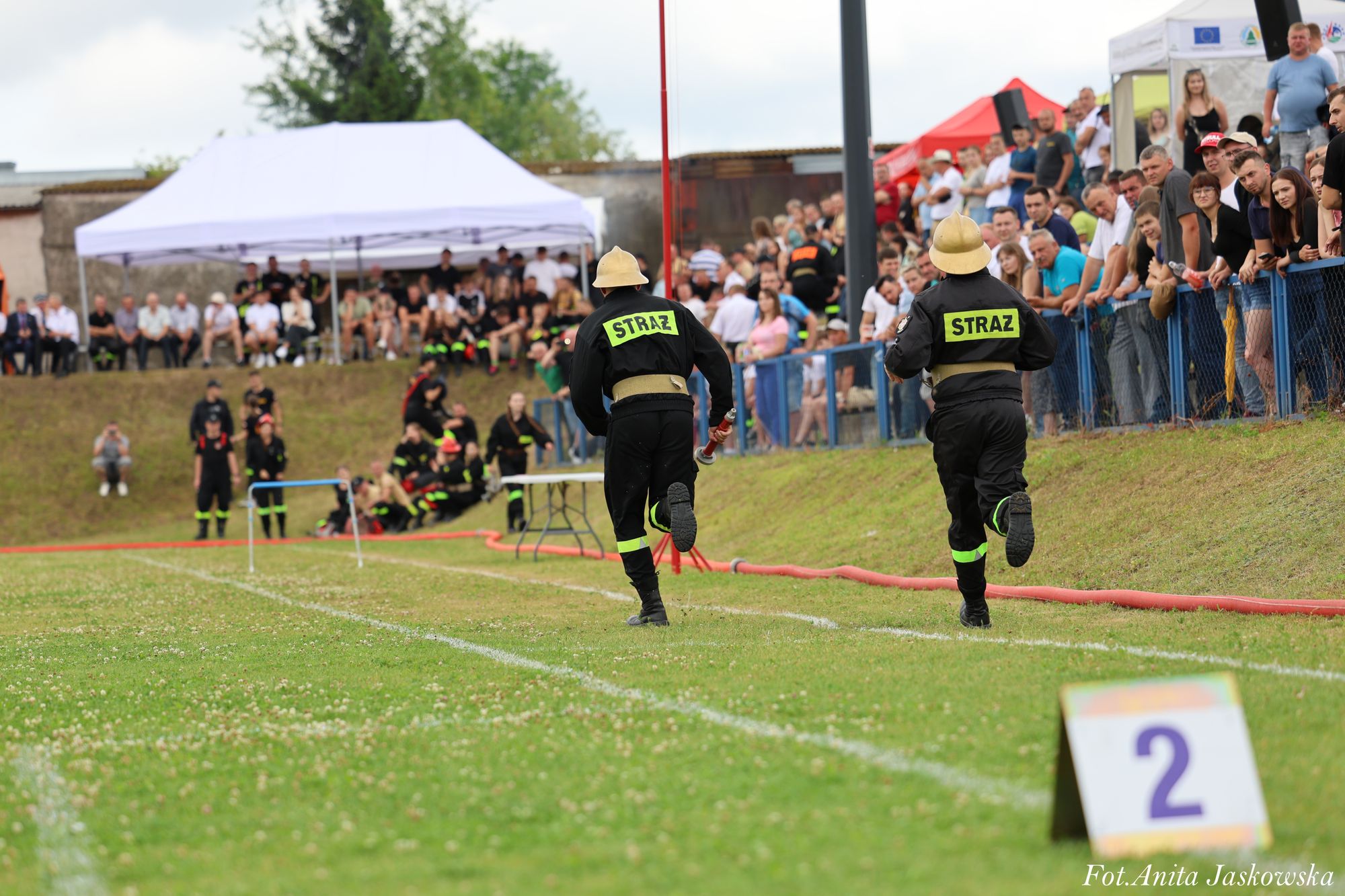 Dwóch strażaków w czarnych mundurach z zielonym napisem "STRAŻ" na plecach biegnie po zielonym boisku, po prawej stronie tłum widzów za niebieską barierką.