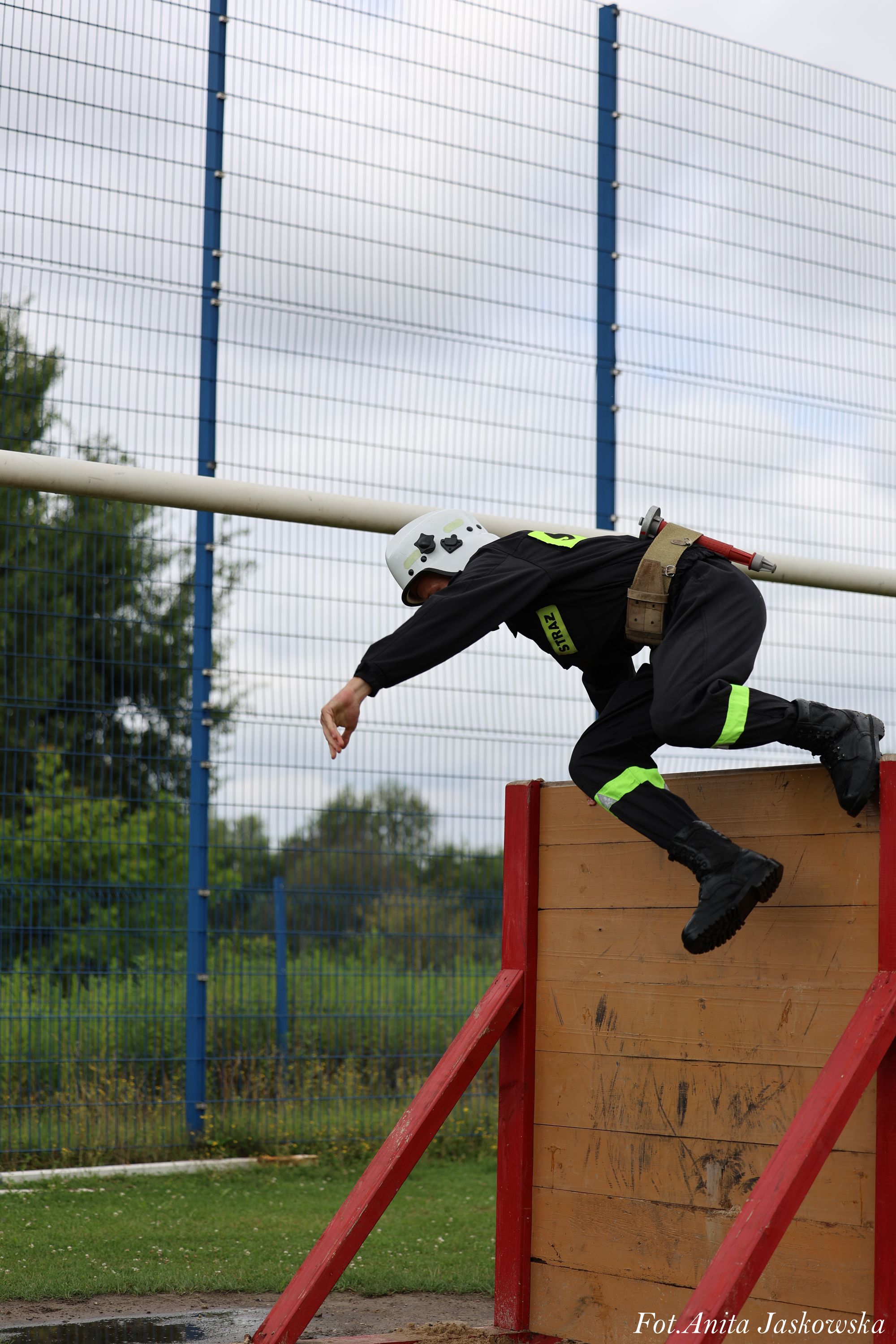 Strażak w czarnym ubraniu z odblaskowymi pasami i białym hełmem pokonujący drewnianą przeszkodę z czerwonymi podporami na tle siatki i zieleni.
