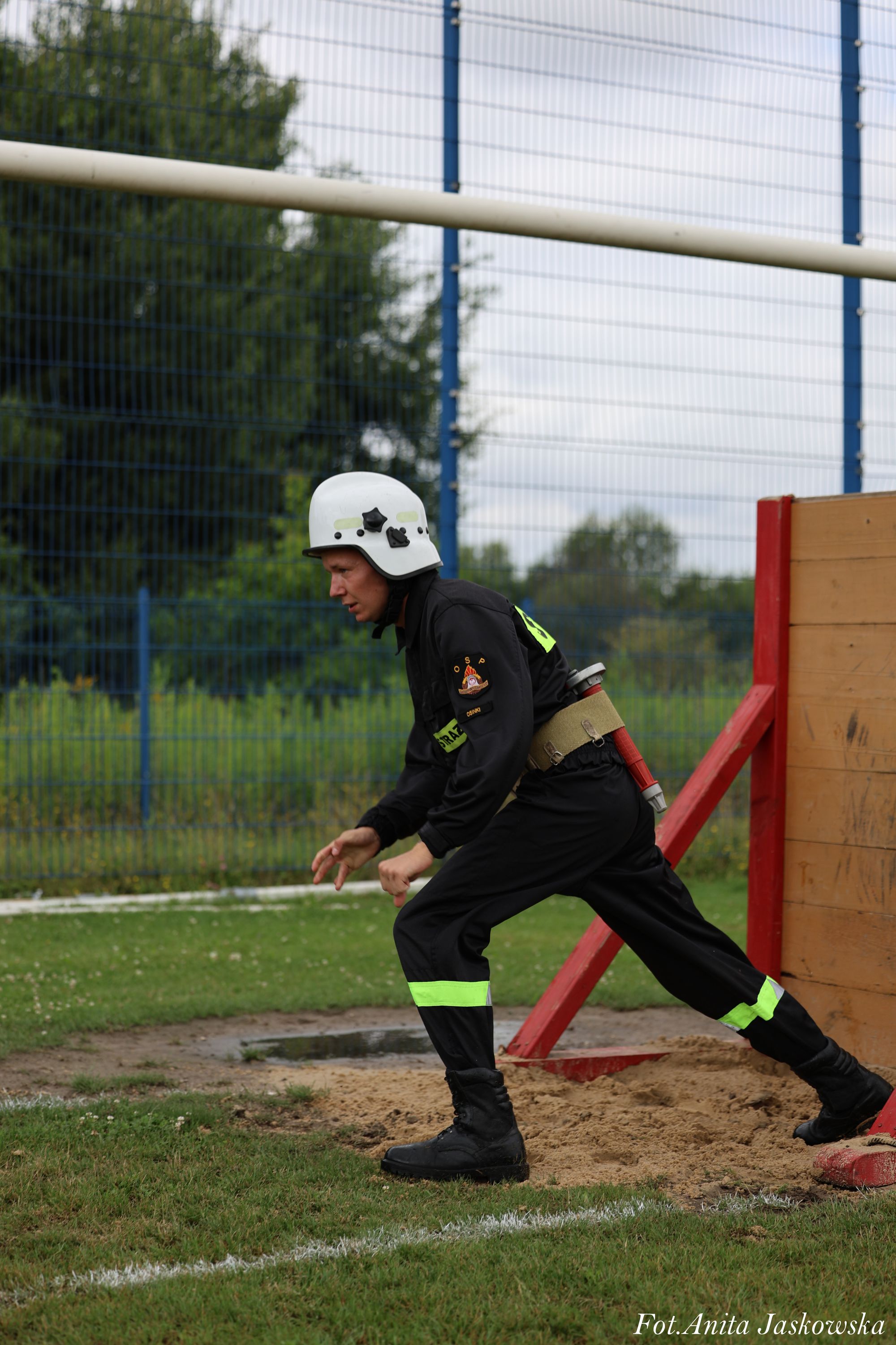 Mężczyzna w czarnym ubraniu strażaka z odblaskowymi paskami i białym kaskiem, stojący na trawie obok drewnianej przeszkody z czerwonymi podporami.