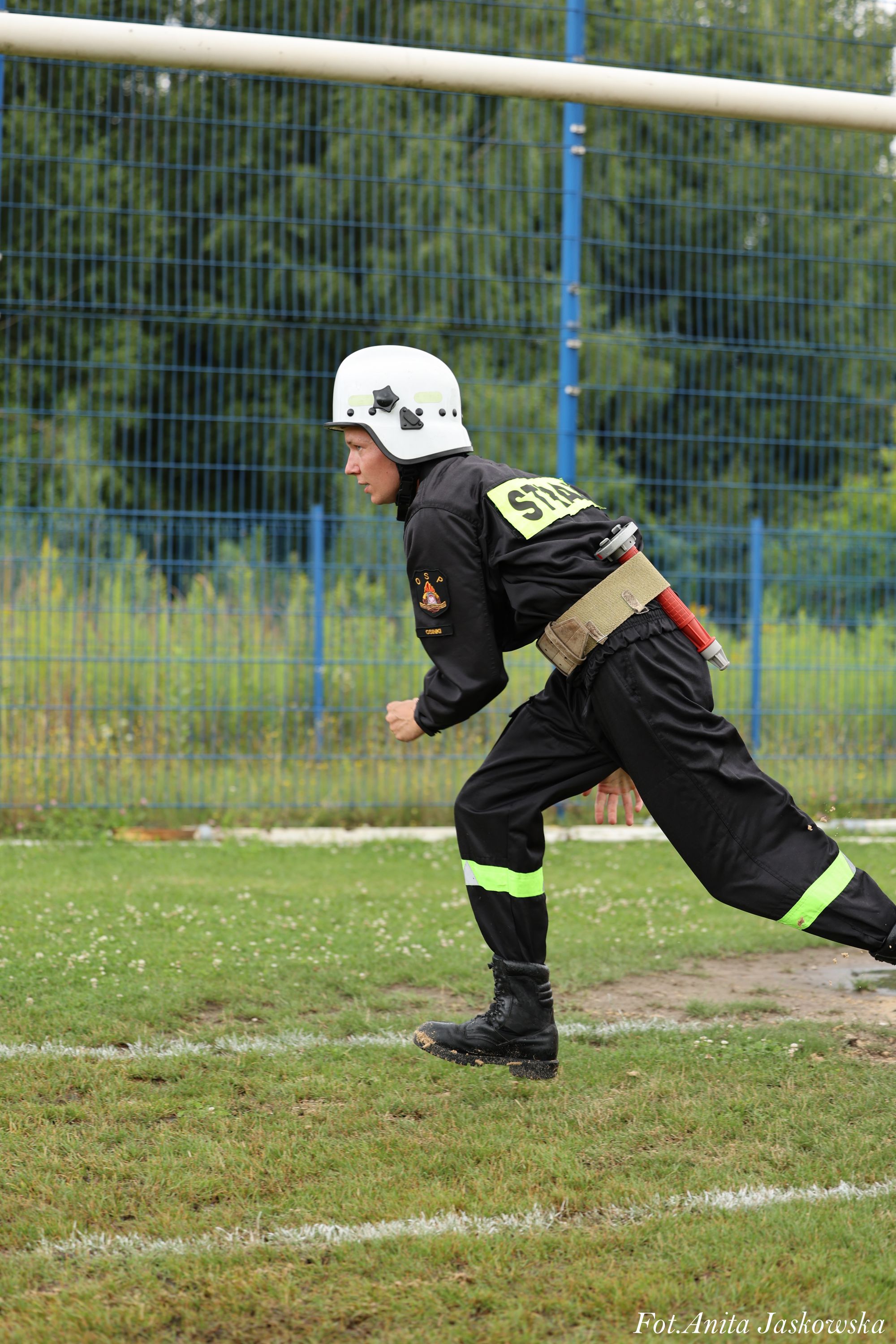 Strażak w czarnym ubraniu z odblaskowymi pasami i białym hełmem biegnie po zielonym boisku z białymi liniami, w tle niebieski metalowy płot i zieleń.