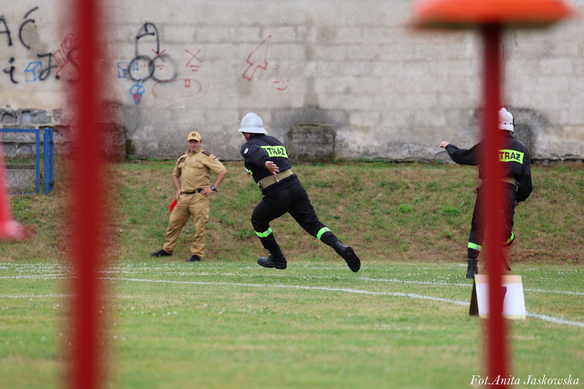 Dwóch strażaków w czarnych uniformach z napisem "STRAŻ" biegnie po zielonym trawniku, w tle mężczyzna w beżowym mundurze stoi na tle ściany z graffiti.