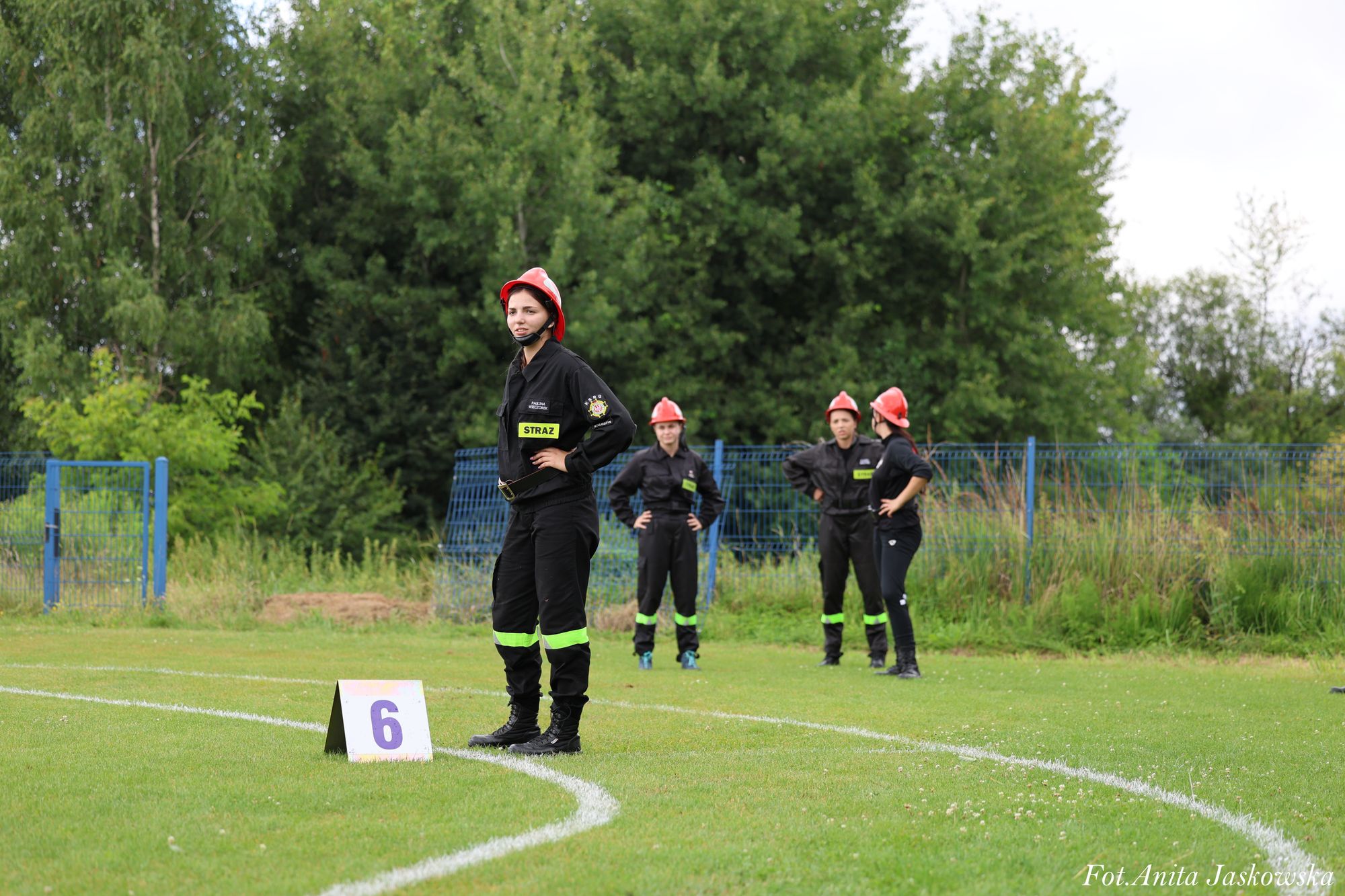 Cztery osoby w czarnych mundurach z odblaskowymi paskami i czerwonych hełmach stoją na zielonej trawie przy białych liniach boiska, obok tabliczka z numerem 6.