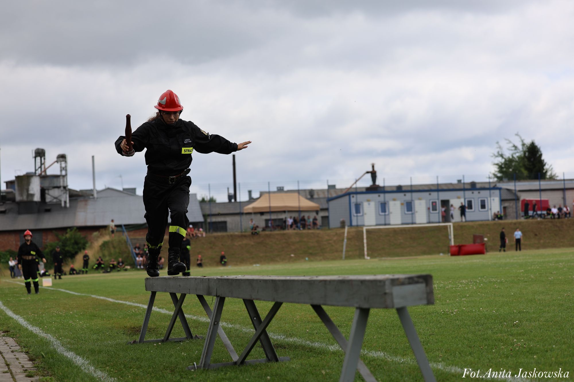 Kobieta w czarnym mundurze i czerwonym kasku biegnie po drewnianej równoważni na zielonym boisku sportowym, trzymając drewniany przedmiot.