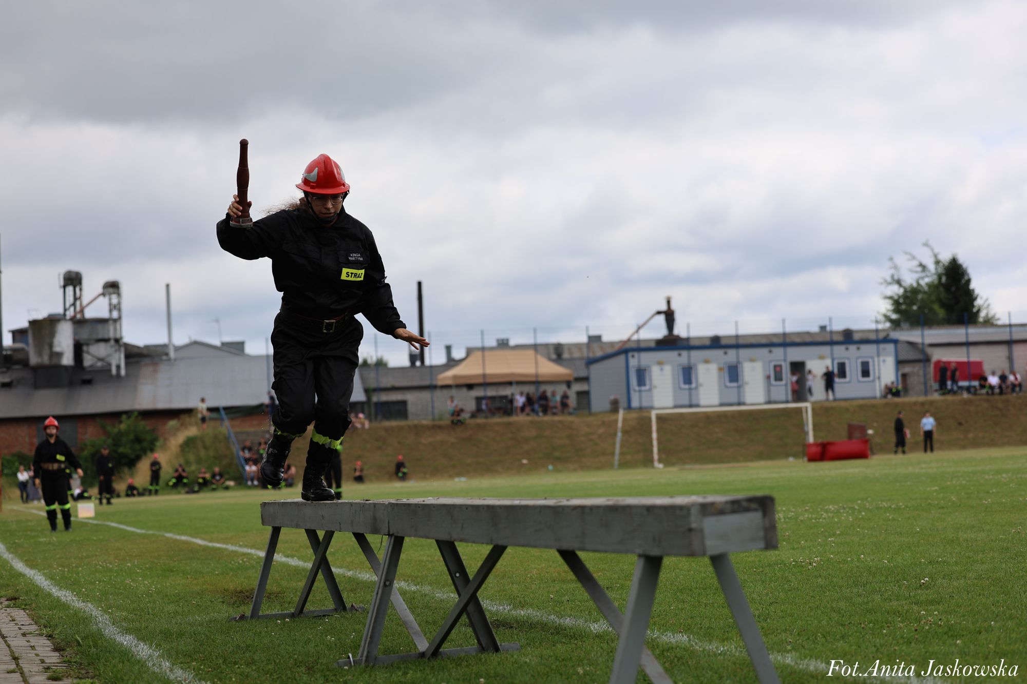 Strażak w czarnym uniformie i czerwonym hełmie biegnie po drewnianej, podwyższonej równoważni na zielonym boisku sportowym pod pochmurnym niebem.