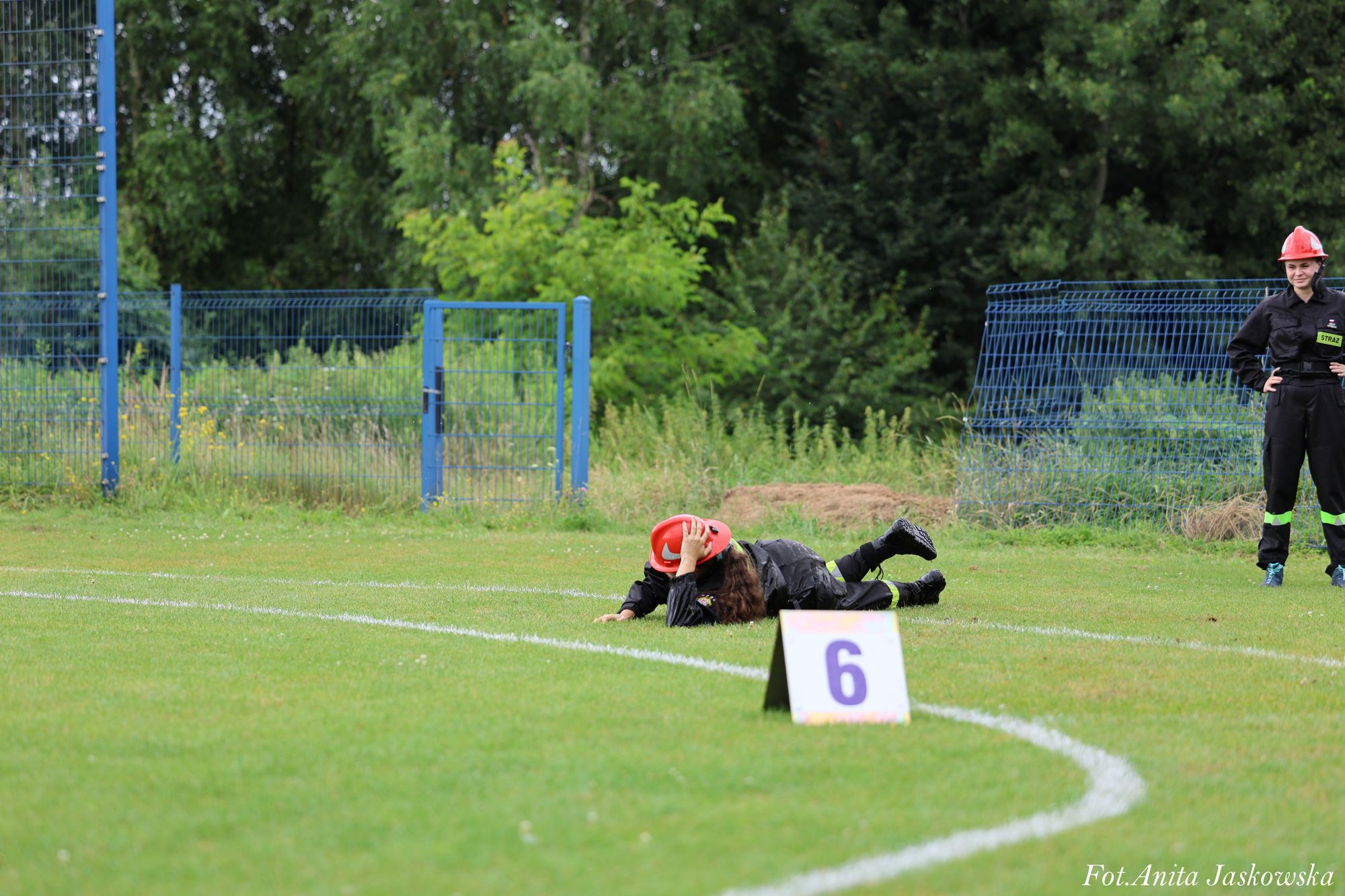Dwie osoby w czarnych mundurach i czerwonych hełmach na zielonej trawie, jedna leży na ziemi, druga stoi obok metalowego ogrodzenia. Przed nimi tablica z cyfrą 6.