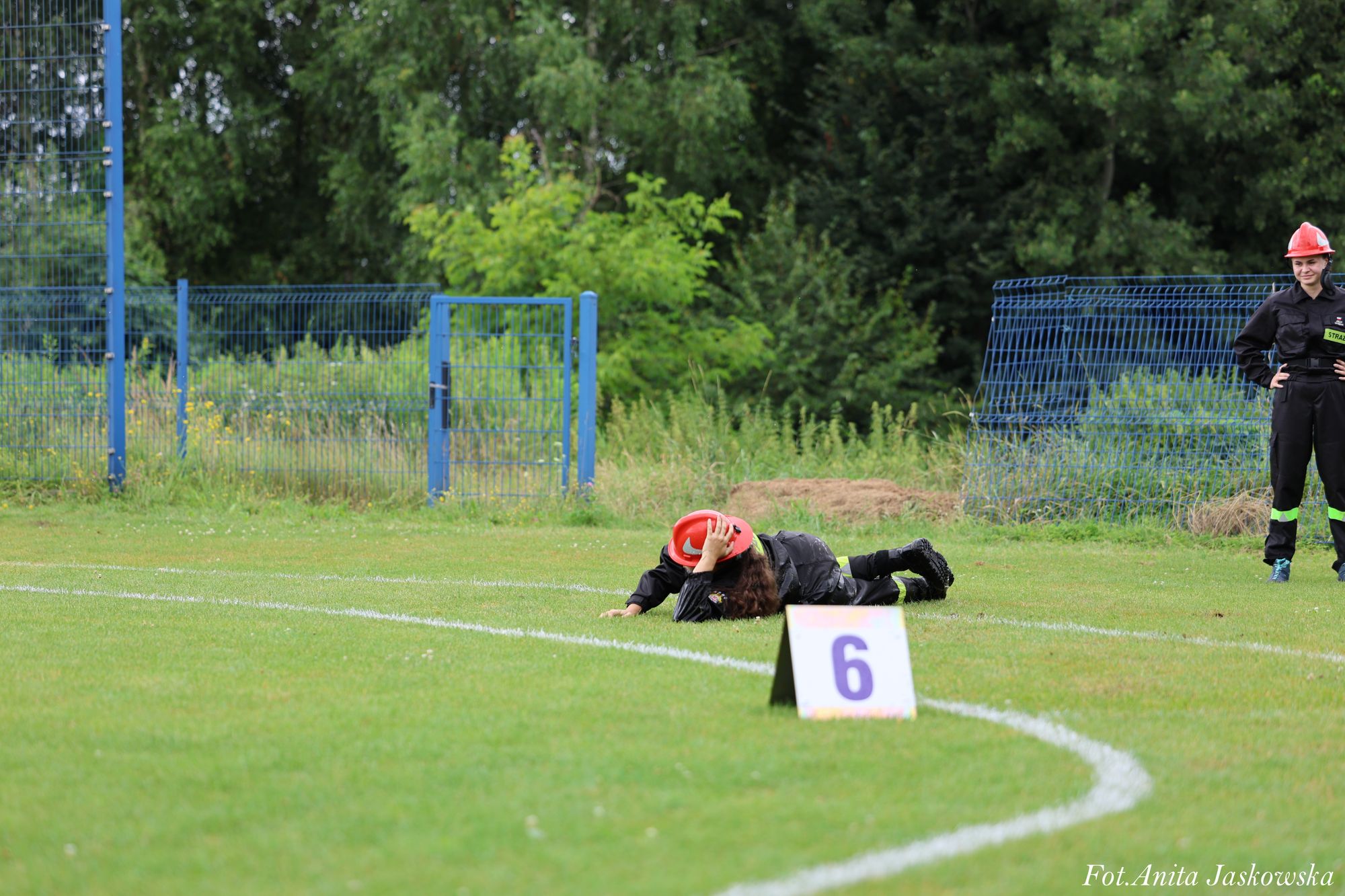 Osoba w czarnym uniformie i czerwonym kasku leży na trawie, druga stoi obok, za nimi niebieskie ogrodzenie i zielone drzewa. Na pierwszym planie tablica z cyfrą 6.