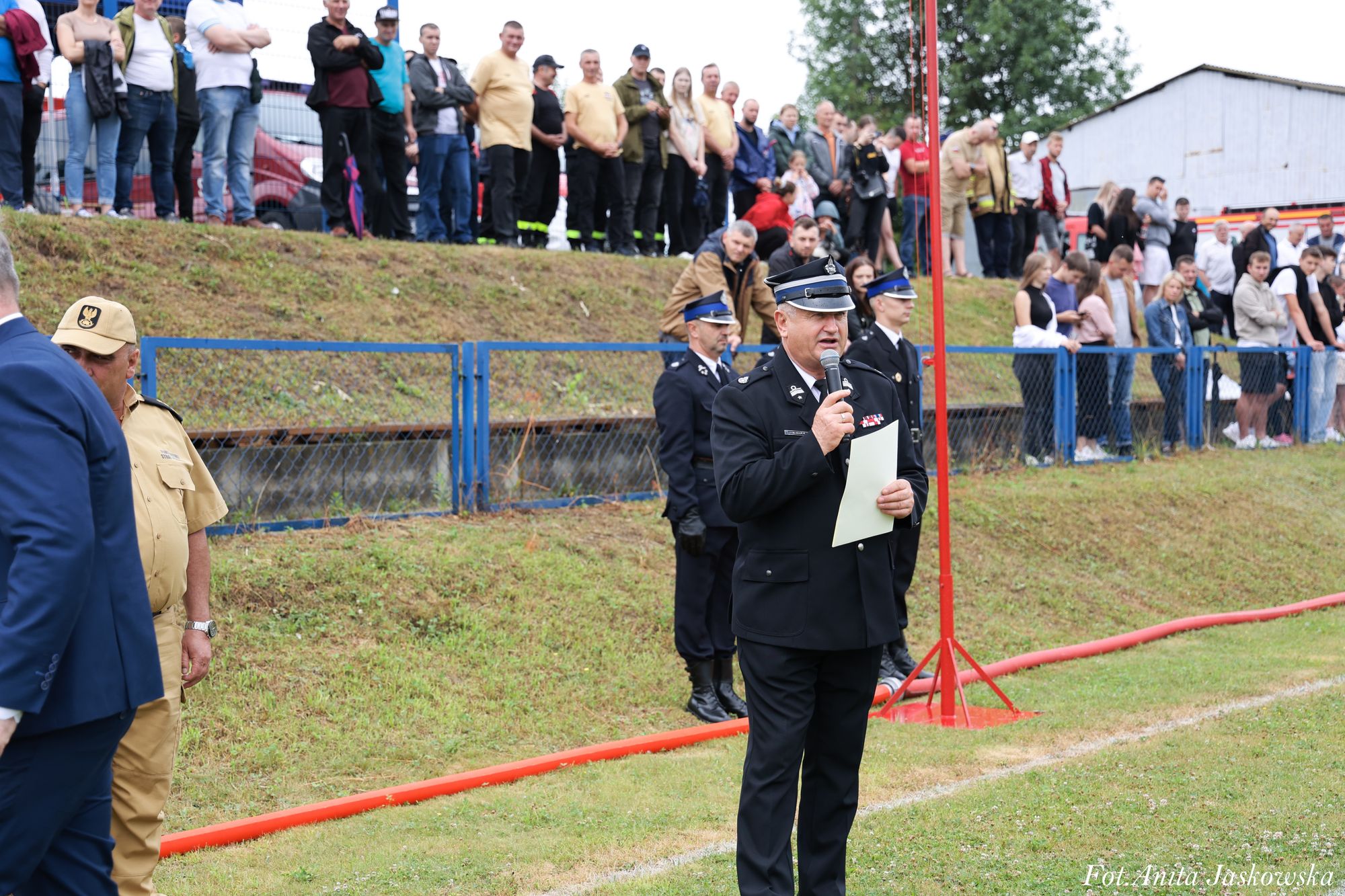 Mężczyzna w czarnym mundurze strażackim z mikrofonem i kartką na tle ludzi stojących na skarpie za niebieskim ogrodzeniem, trawa i czerwony wąż strażacki.