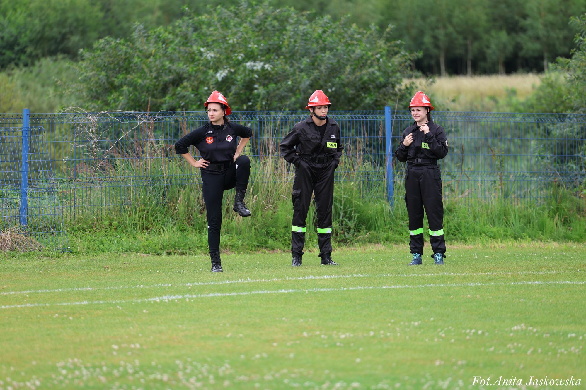 Trzy osoby w czarnych mundurach i czerwonych hełmach stoją na zielonej trawie na tle niebieskiego metalowego ogrodzenia oraz zielonych krzewów i drzew.