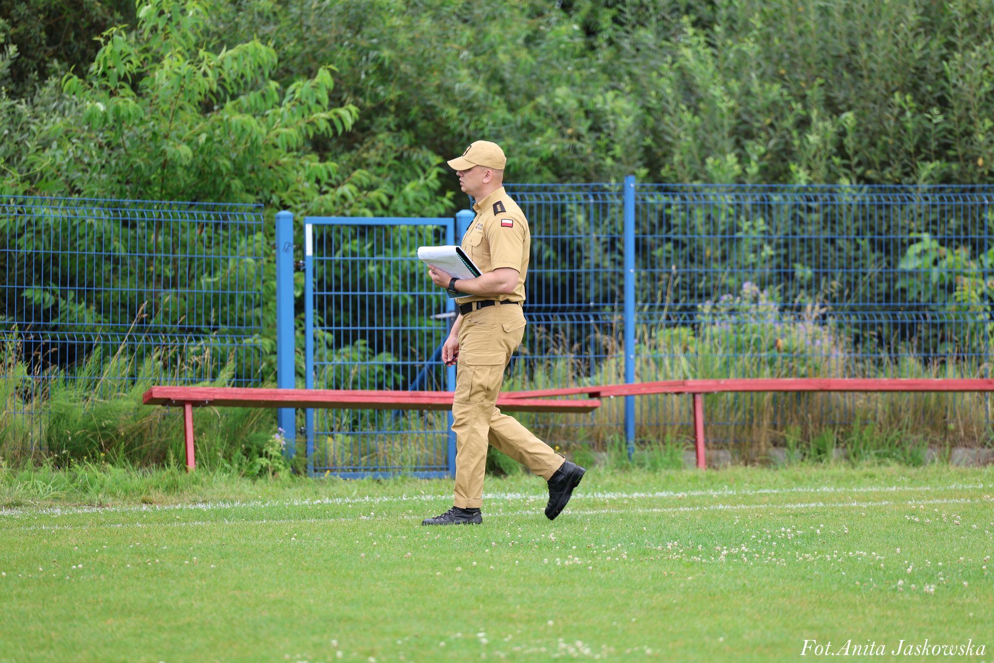 Mężczyzna w beżowym mundurze z czapką i notatnikiem, idący po trawiastej powierzchni, w tle niebieskie ogrodzenie i zielone krzewy.