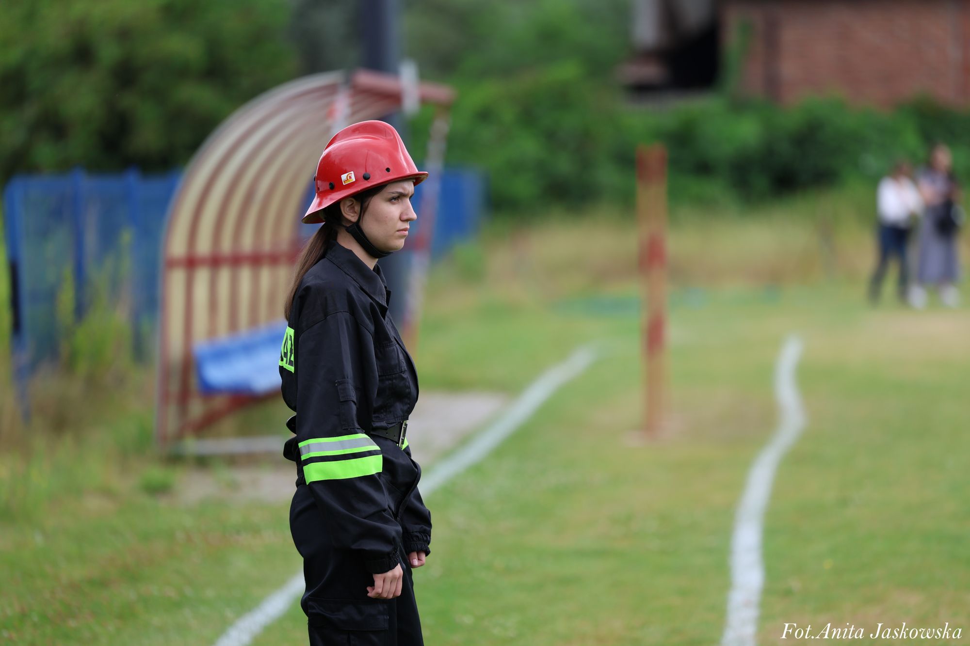 Kobieta w czarnym ubraniu strażackim z neonowożółtymi paskami i czerwonym hełmem stoi na trawie obok boiska sportowego z białymi liniami.