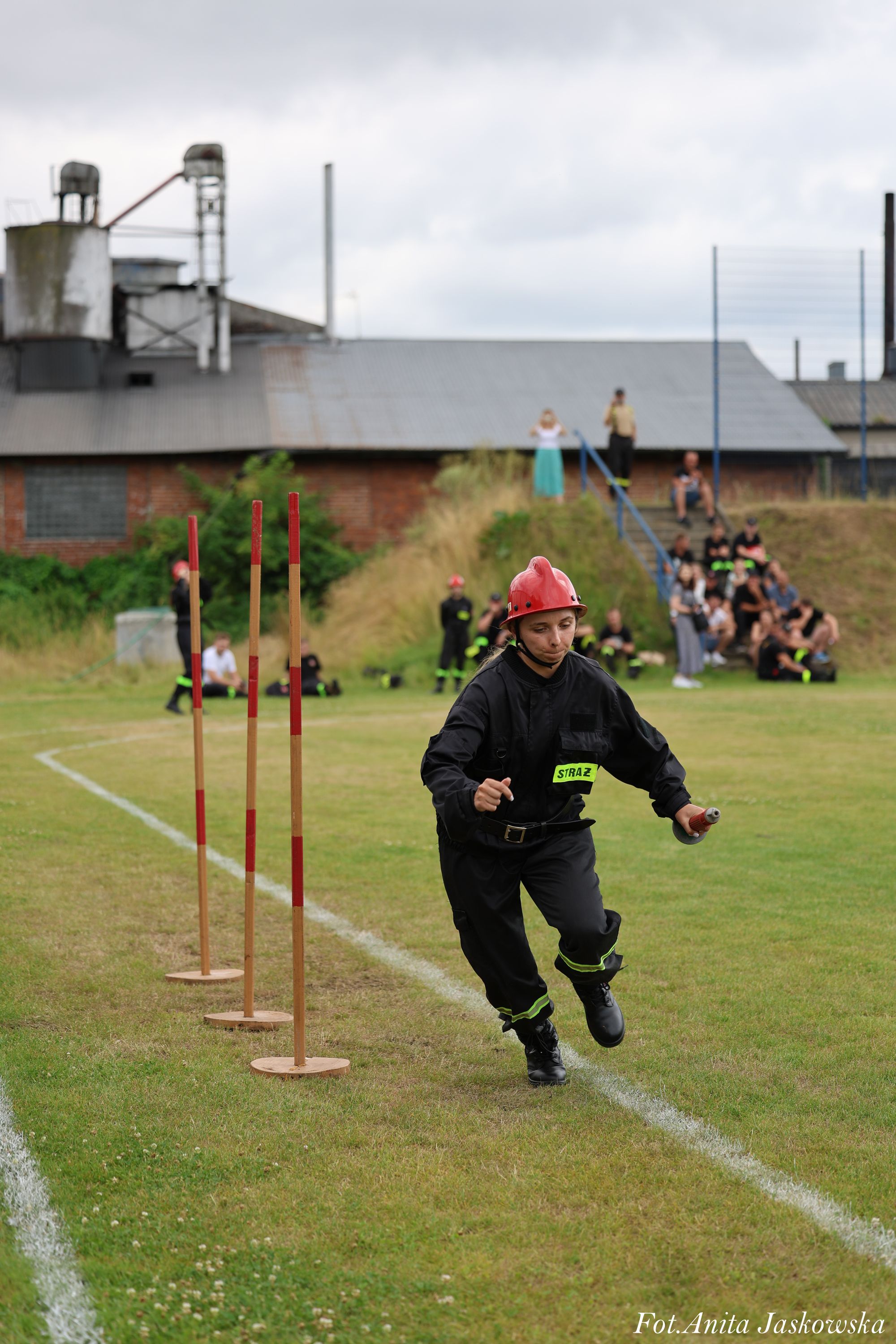Kobieta w czarnym ubraniu strażackim i czerwonym kasku biegnie po trawiastej murawie obok trzech pionowych słupków z czerwonymi pasami. W tle ludzie na trawie i schodach.