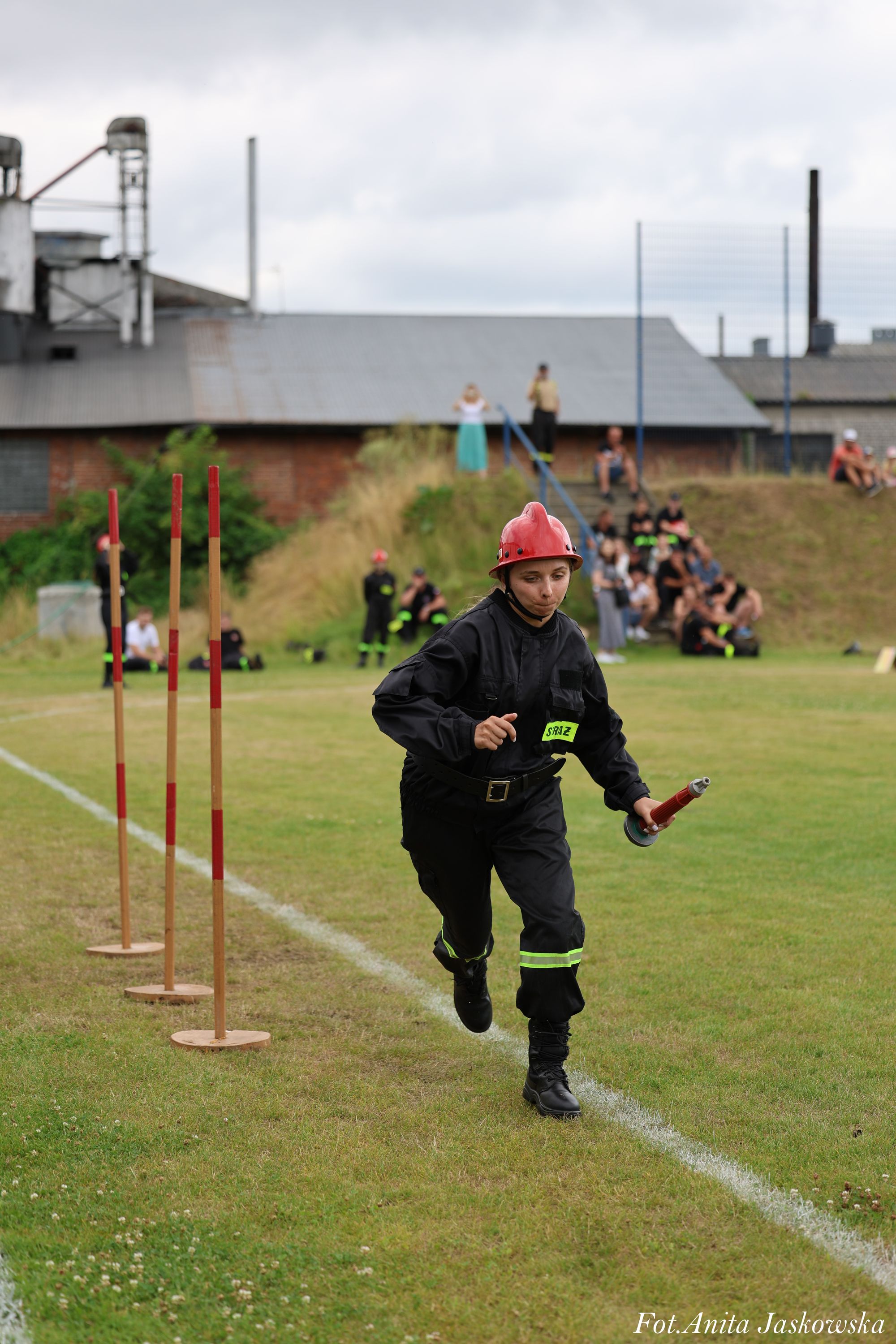 Osoba w czarnym stroju strażackim i czerwonym hełmie biegnie po trawie obok trzech pionowych tyczek z czerwonymi paskami, w tle ludzie na wzgórzu i budynki przemysłowe.