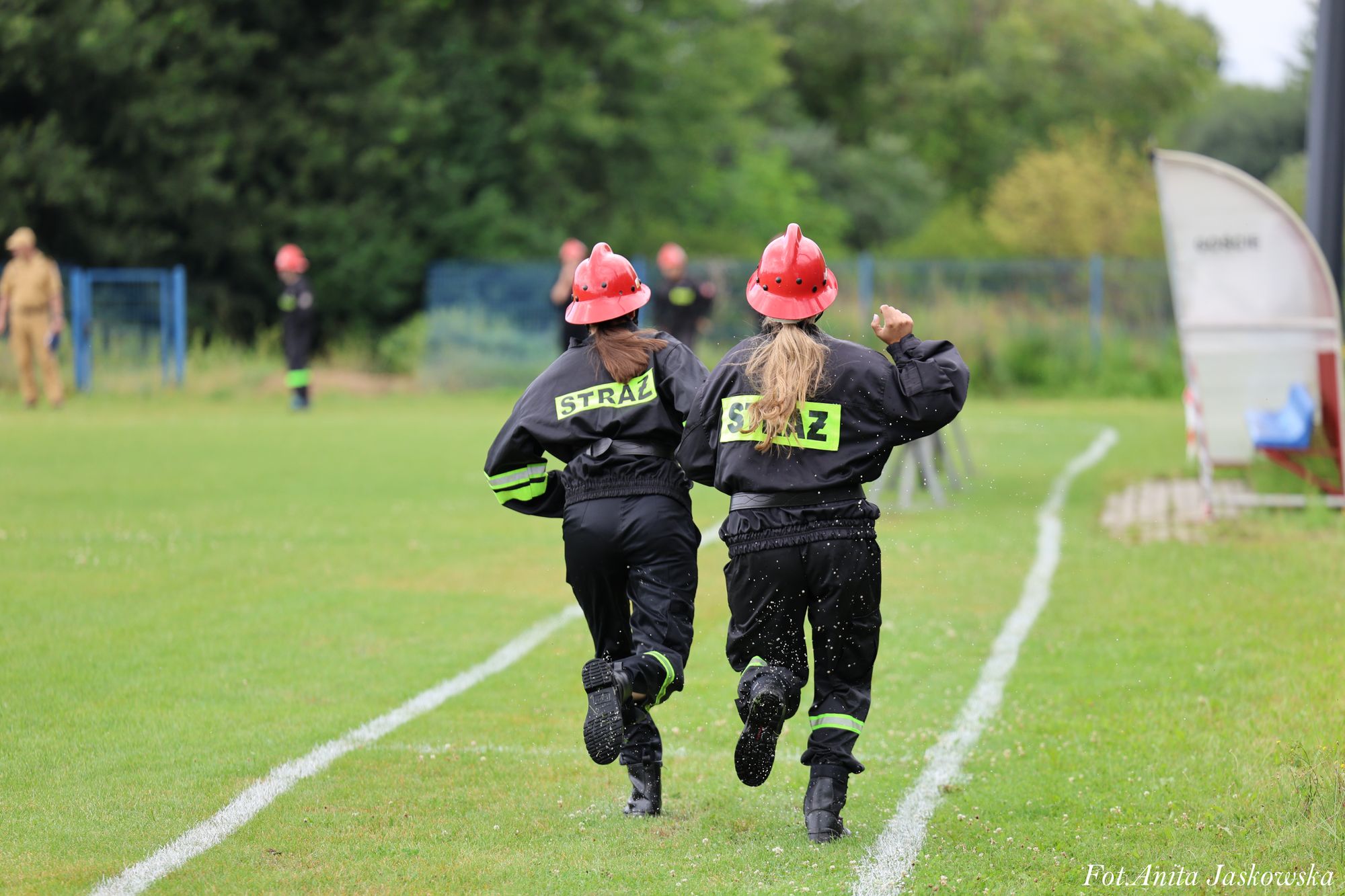 Dwie kobiety w czarnych mundurach strażackich z odblaskowymi napisami STRAŻ i czerwonych hełmach biegną po zielonym boisku z białymi liniami, w tle drzewa i rozmyci ludzie.