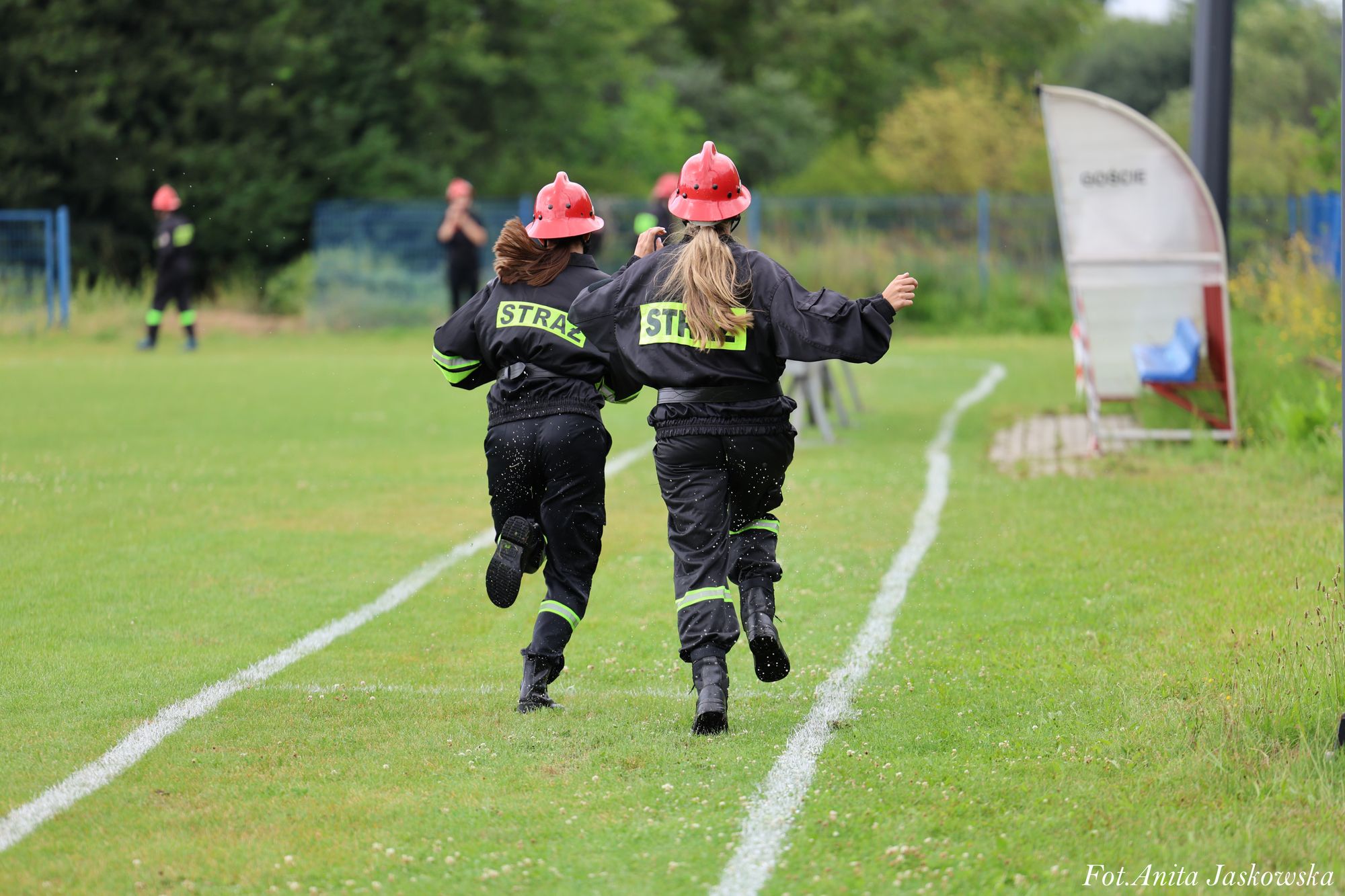 Dwie osoby w czarnych mundurach strażackich z czerwonymi hełmami biegną po zielonym boisku z białymi liniami, w tle drzewa i nieostre sylwetki osób.