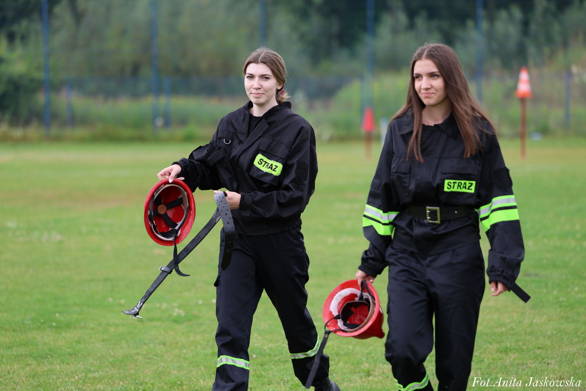 Dwie młode kobiety w czarnych mundurach strażackich z odblaskowymi paskami, każda trzyma czerwoną hełm strażacki, stoją na zielonej trawie.