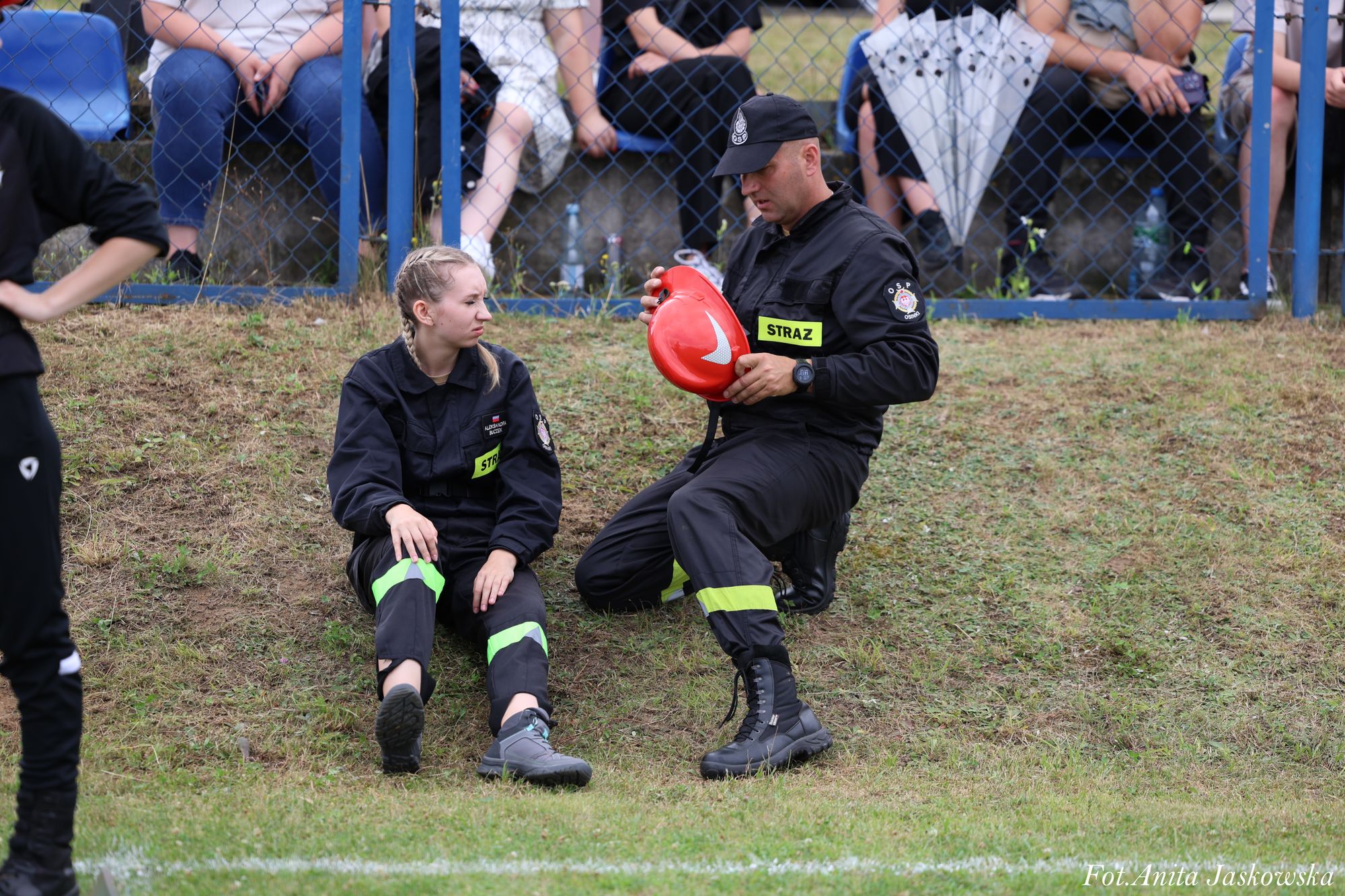 Dwóch strażaków w czarnych mundurach z żółtymi odblaskami, mężczyzna trzyma czerwony hełm, kobieta siedzi na trawie, w tle siatka i osoby siedzące na murku.
