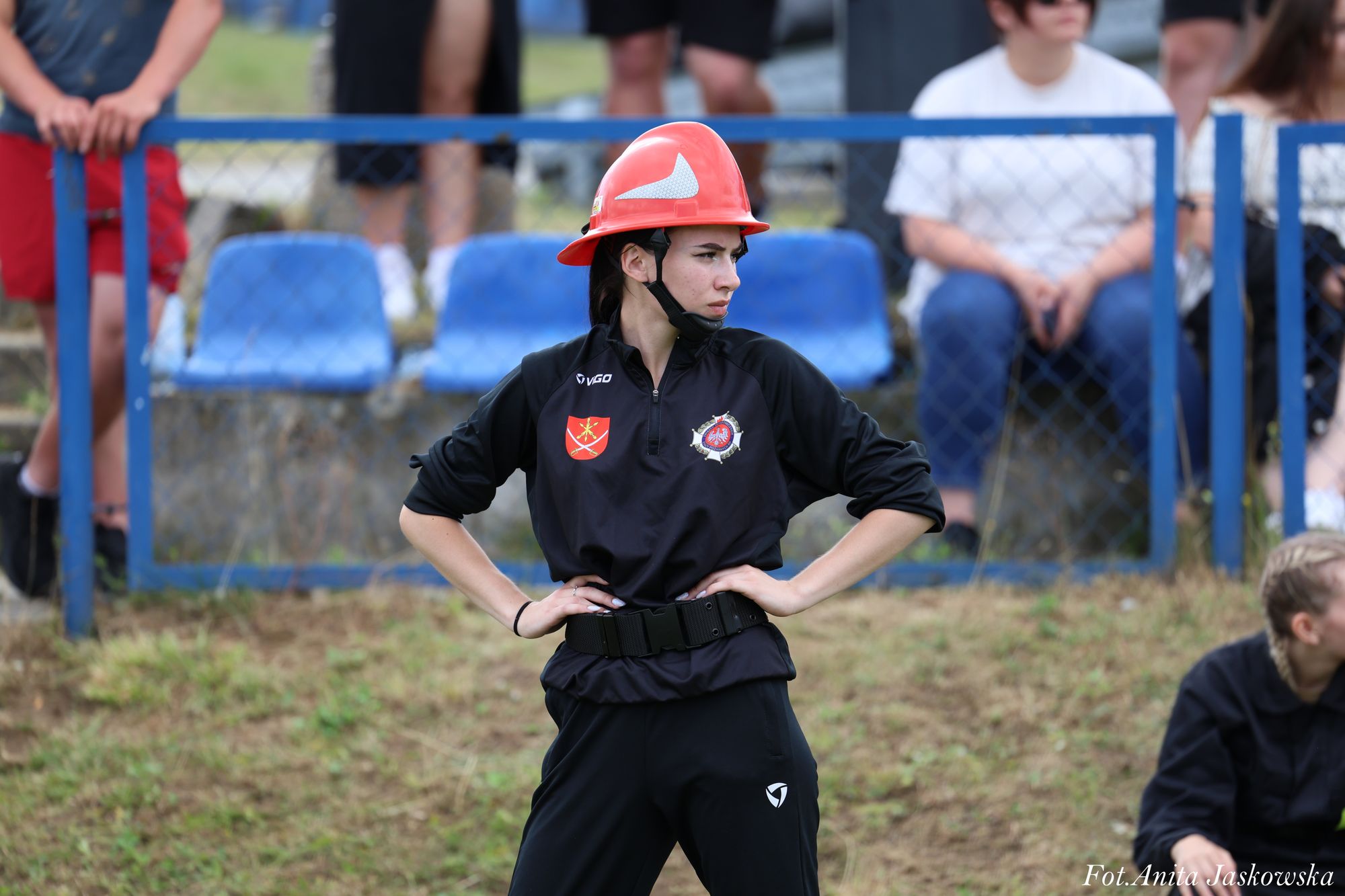 Kobieta w czerwonym kasku i czarnym stroju sportowym stoi na tle niebieskich ławek i ludzi za ogrodzeniem z siatki.